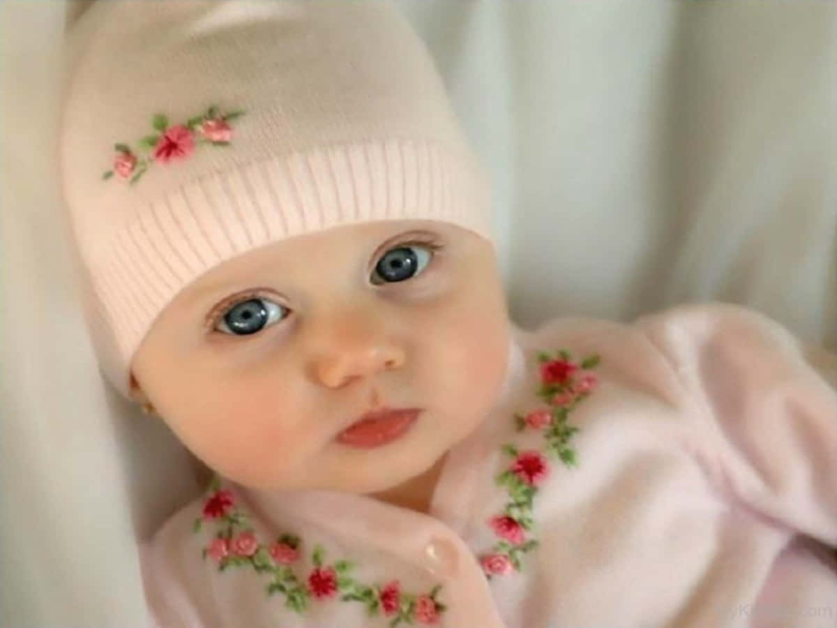 Baby Girl 桌布 壁紙 — 一個擁有引人注目藍色眼睛的嬰兒特寫，戴著柔軟的粉紅色針織帽和搭配的服裝，飾有精緻的花卉刺繡。