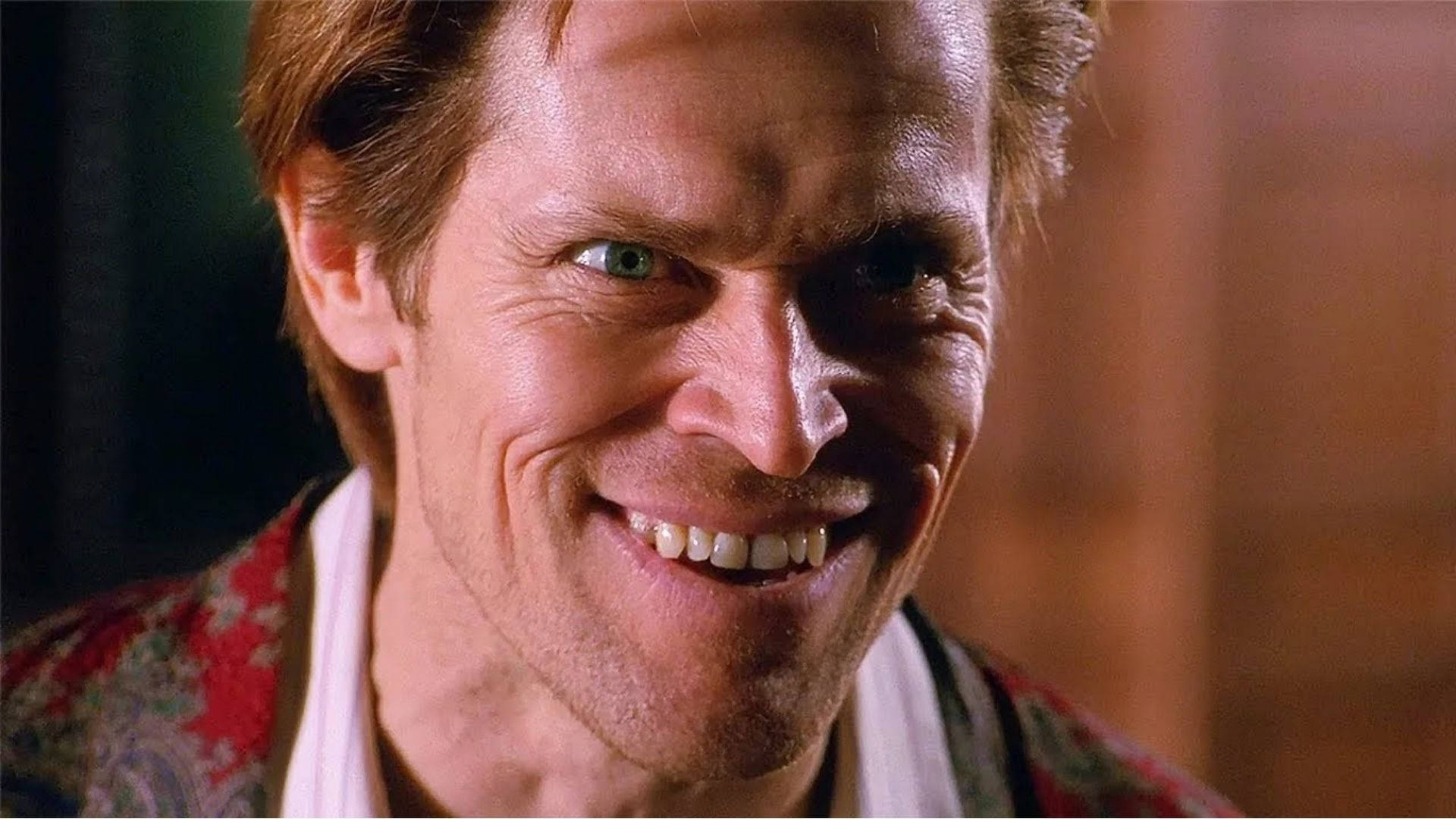 Willem Dafoe 桌布 壁紙 — 一個擁有強烈綠色眼睛和寬闊露齒笑容的男人，穿著圖案紅色長袍，背景溫暖模糊。