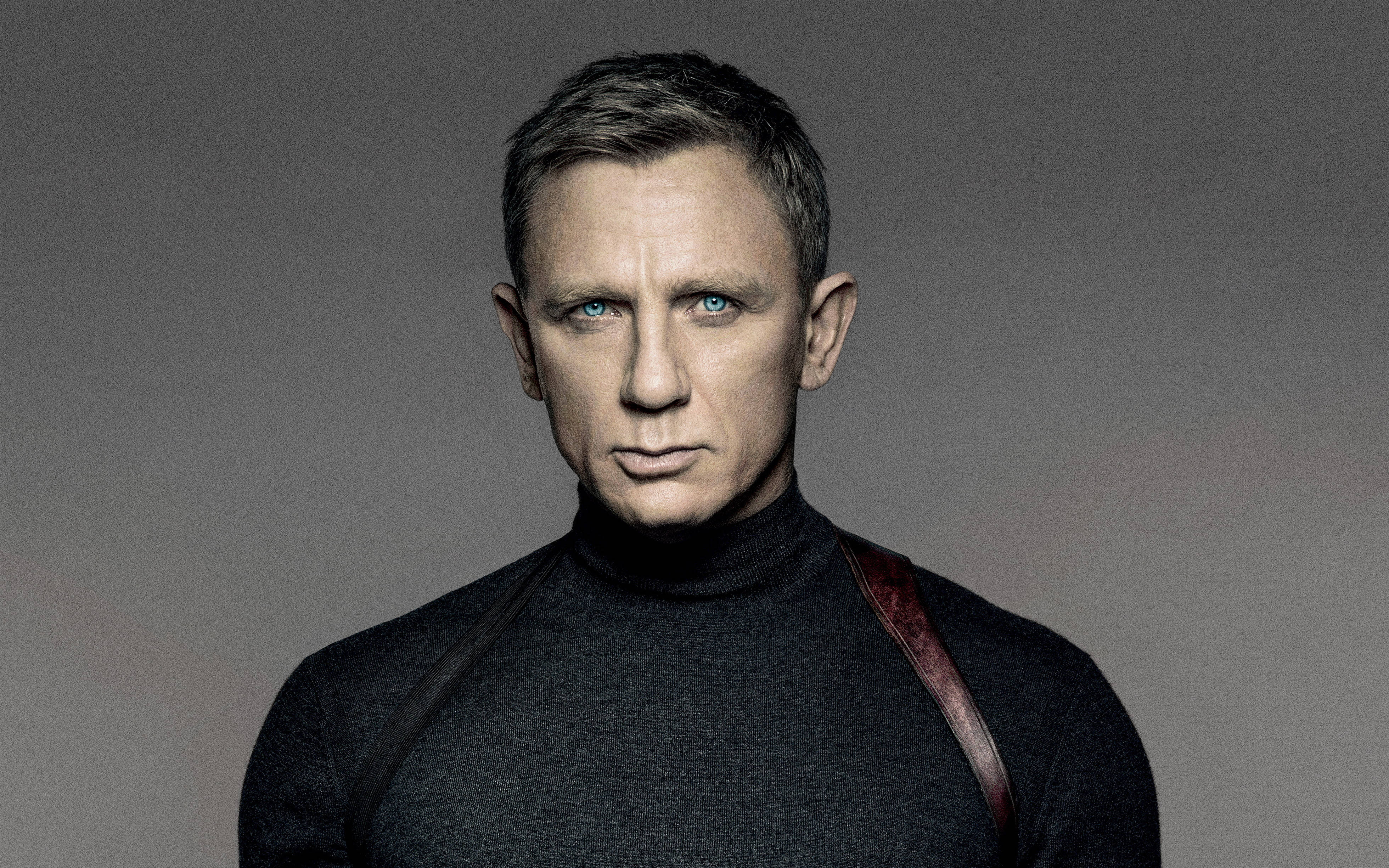 Daniel Craig 桌布 壁紙 — 一個擁有強烈藍色眼睛和短黑髮的男人直視鏡頭，穿著黑色高領毛衣，肩上掛著一條皮革帶子，背景為灰色。