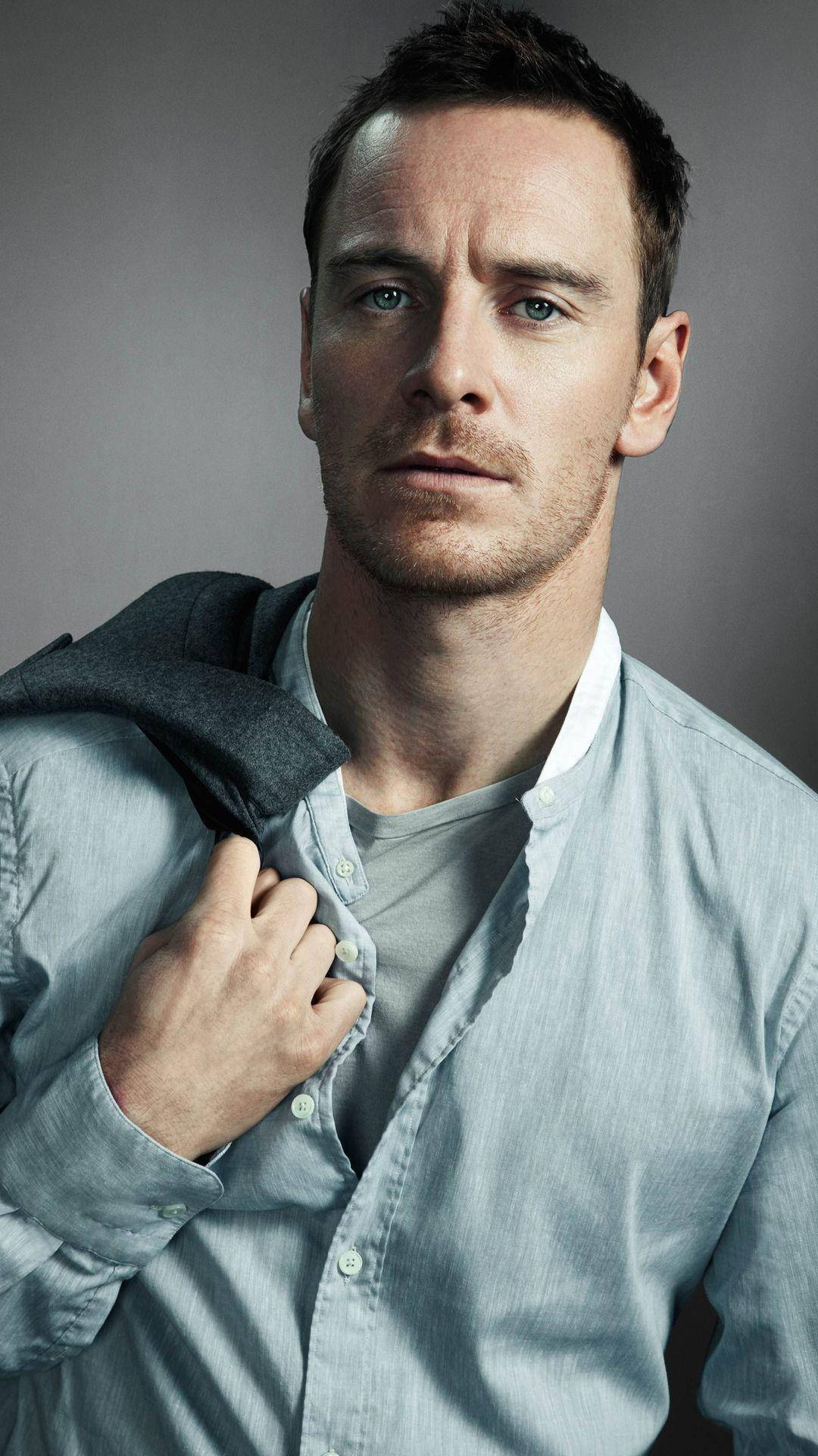 michael 桌布 壁紙 — 一個擁有強烈藍色眼睛和鬍渣的男人，在灰色中性背景前，將一件深色夾克搭在肩上，直視鏡頭。