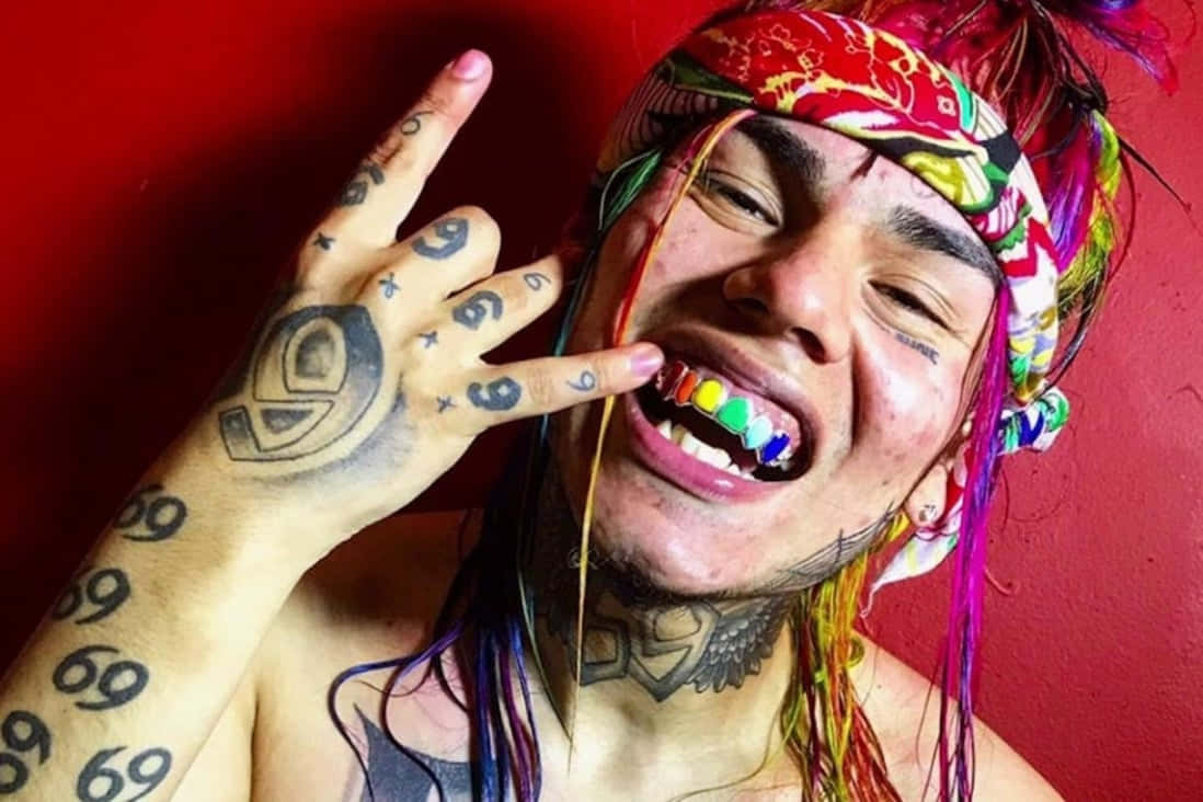69 Rapper 桌布 壁紙 — 一個擁有彩虹色頭髮和彩色牙套的人，在鮮紅色的牆壁前展示有刺青的手。