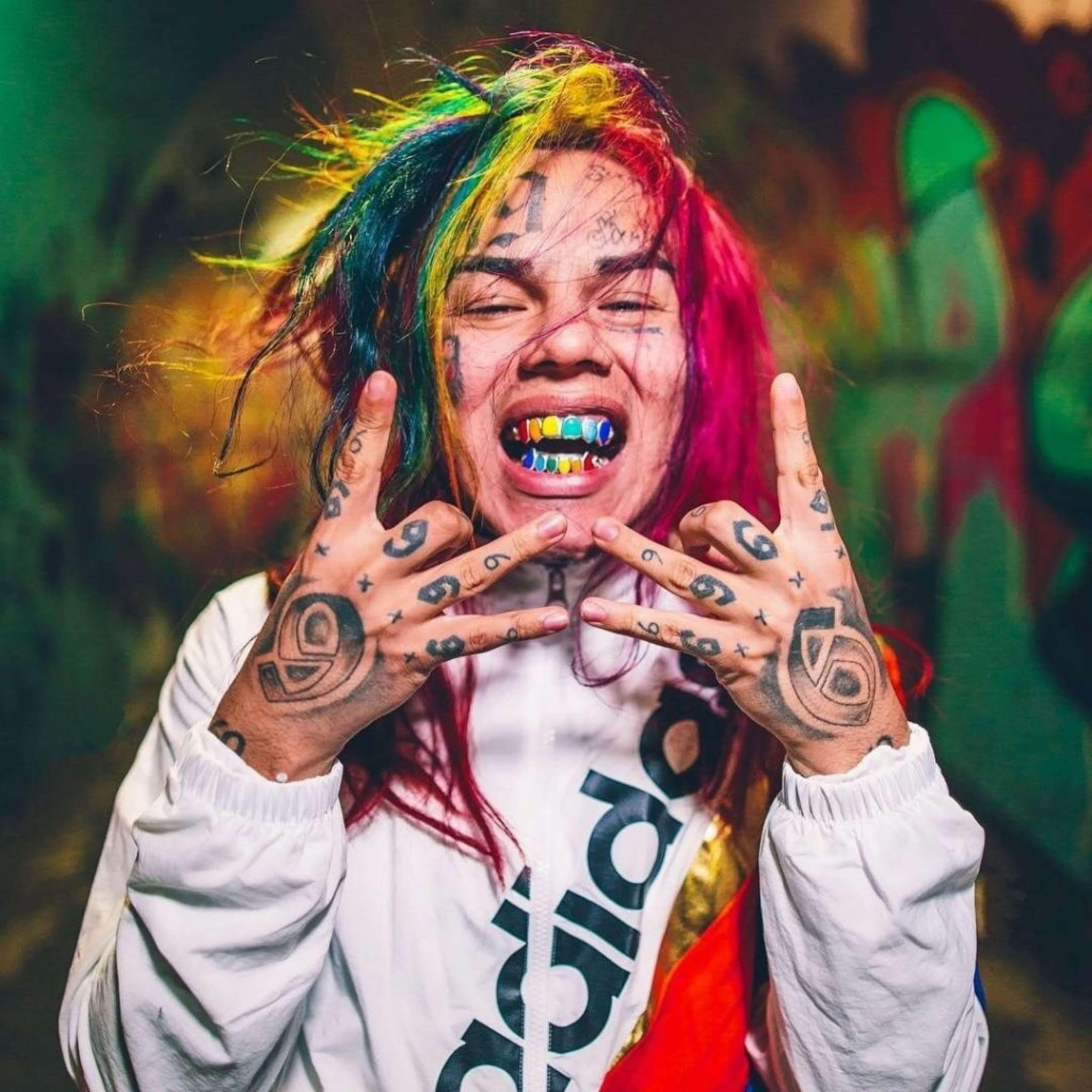 69 Rapper 桌布 壁紙 — 一個擁有彩虹頭髮和刺青的人對著塗鴉牆做出搖滾手勢。