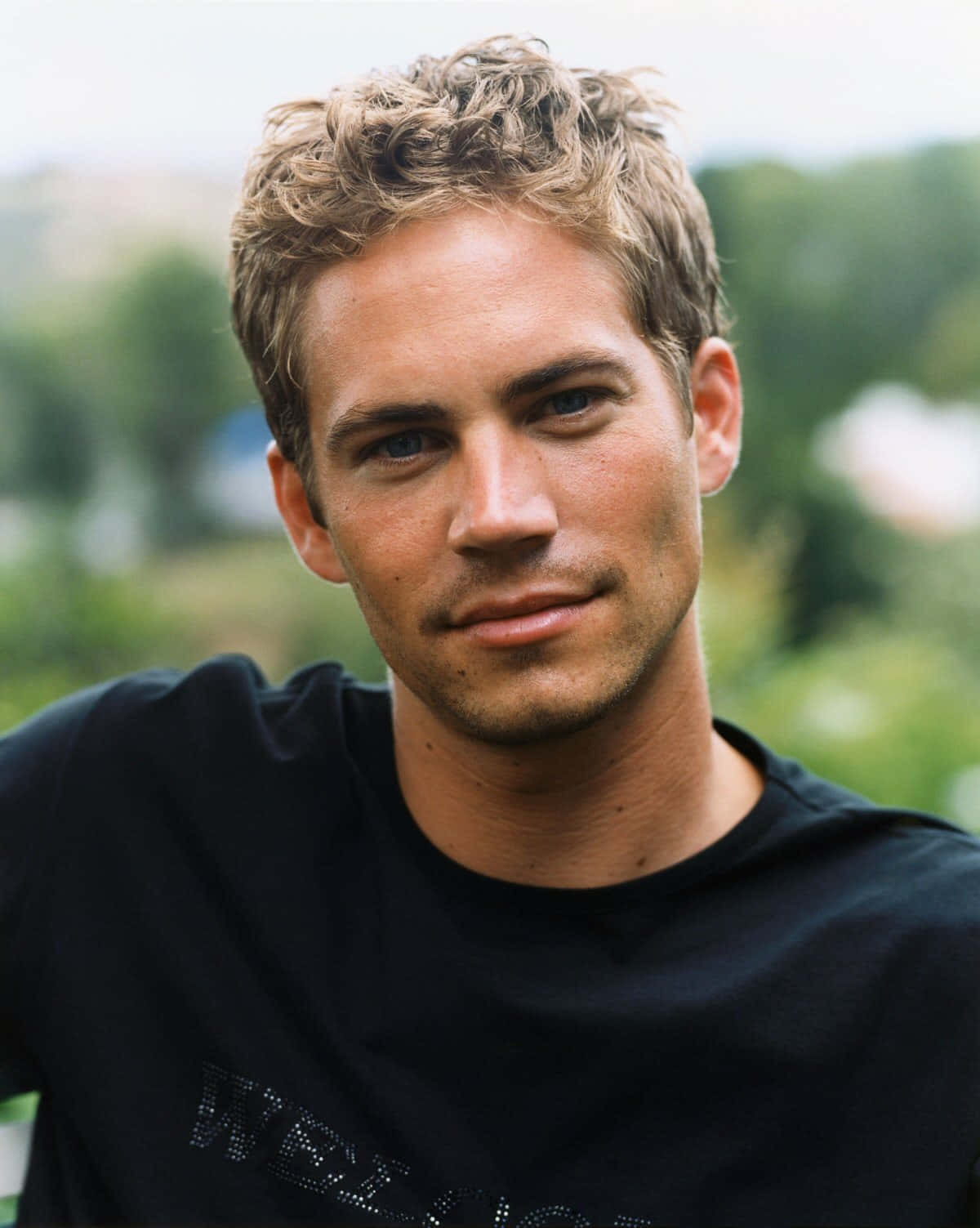 Paul Walker 桌布 壁紙 — 一個擁有捲曲金髮和藍眼睛的男人穿著黑色T恤，以模糊的綠色戶外背景為襯托的特寫肖像。