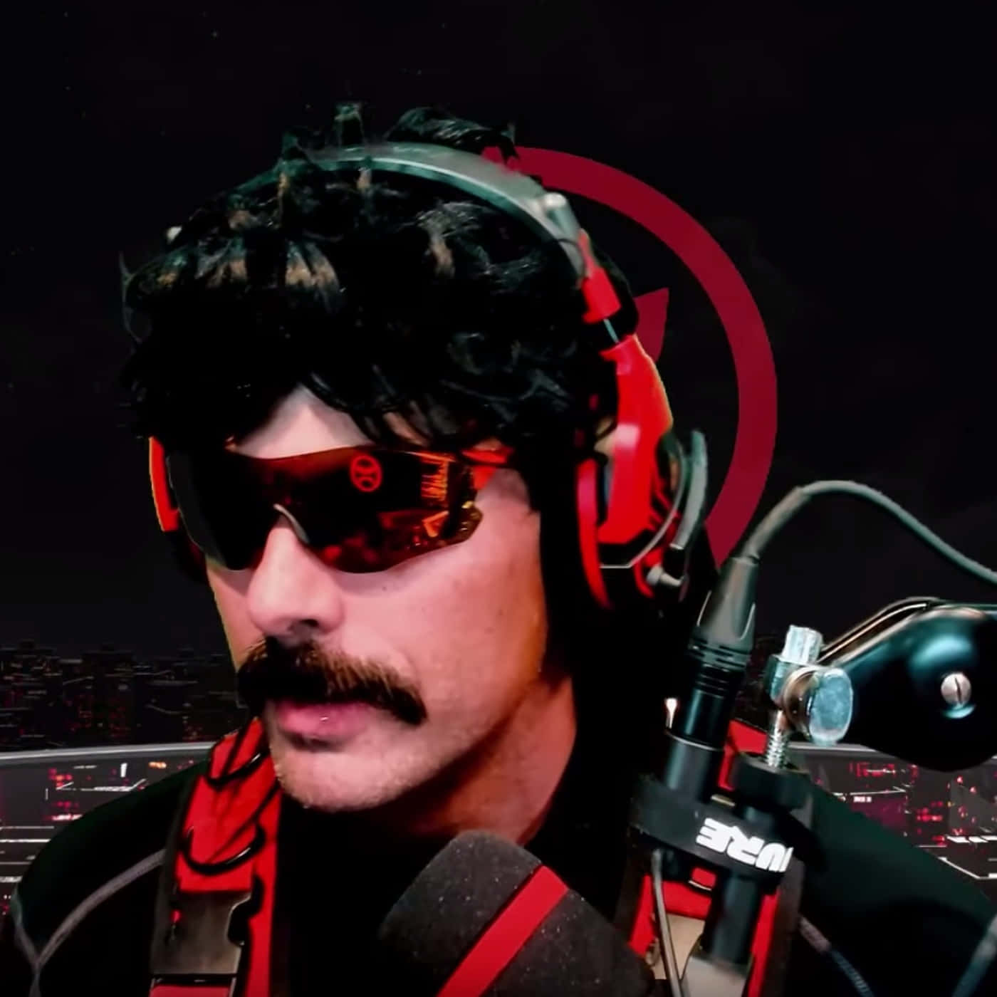Dr Disrespect 桌布 壁紙 — 一個擁有捲髮和太陽眼鏡的男人，戴著紅色耳機，在黑暗的城市背景下對著麥克風說話。