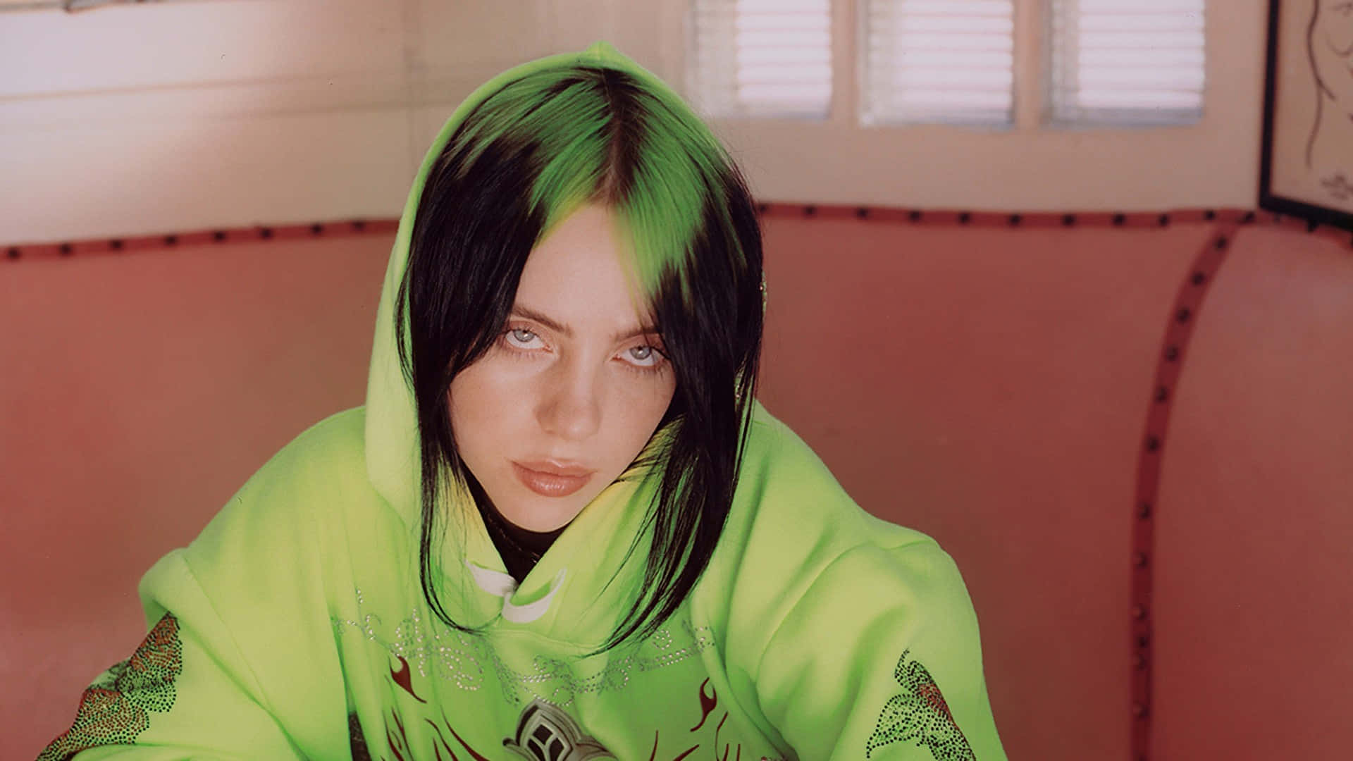 Billie Eilish Laptop 桌布 壁紙 — 一個擁有搶眼綠色和黑色頭髮的人穿著螢光綠色連帽衫，坐在粉紅色沙發上，背景有白色百葉窗。
