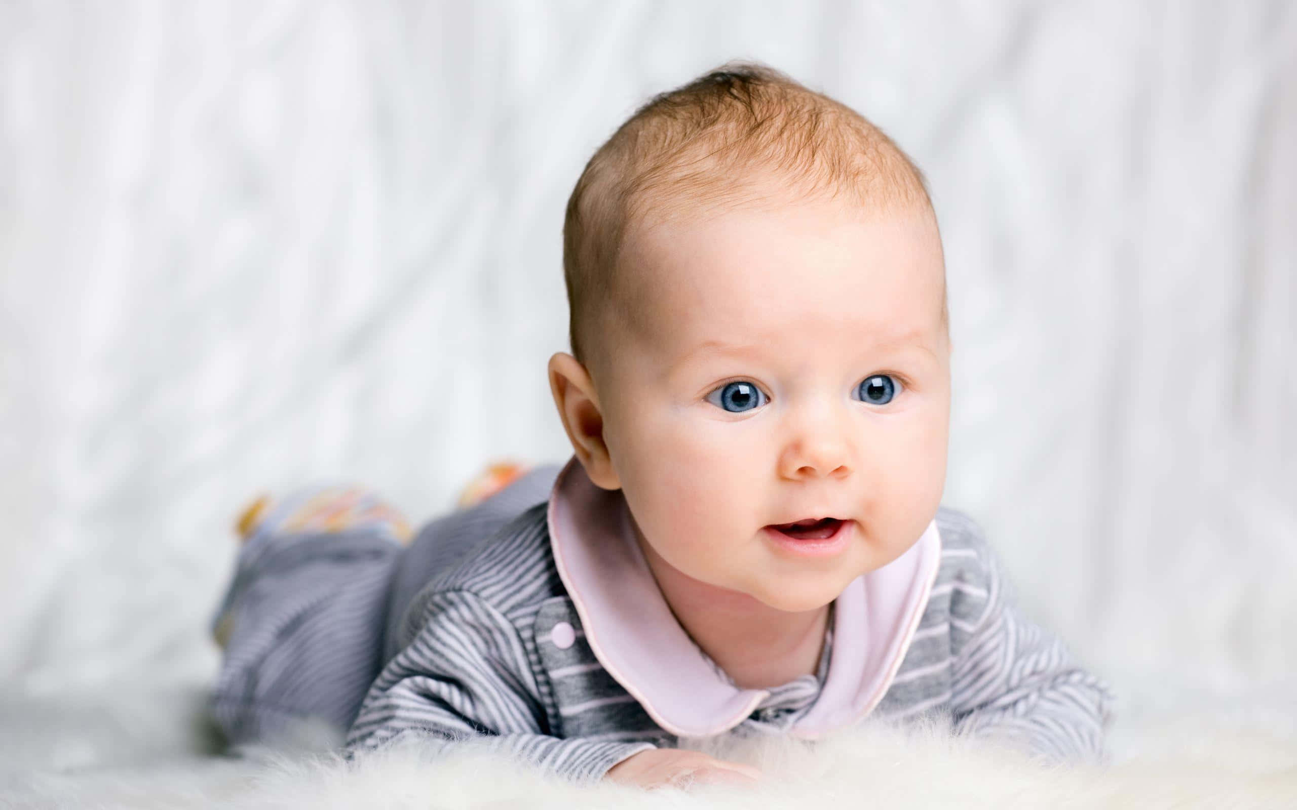 cute baby 桌布 壁紙 — 一個擁有明亮藍眼睛的嬰兒躺在毛絨絨的白色表面上，好奇地看著相機。