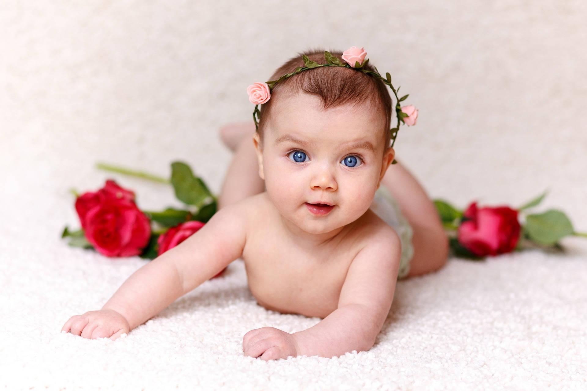 Very Cute Baby 桌布 壁紙 — 一個擁有明亮藍眼睛的嬰兒躺在白色紋理表面上，戴著粉紅色玫瑰皇冠，周圍環繞著紅玫瑰。