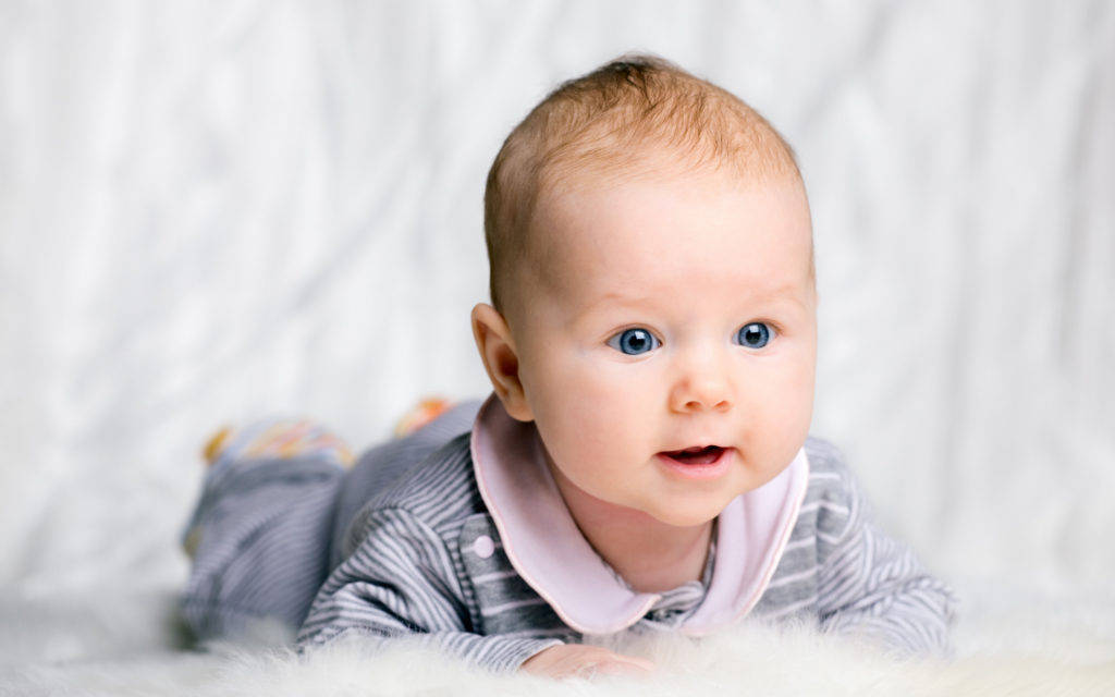 Baby Boy 桌布 壁紙 — 一個擁有明亮藍色眼睛的嬰兒躺在柔軟的白色紋理表面上，好奇地看著相機。