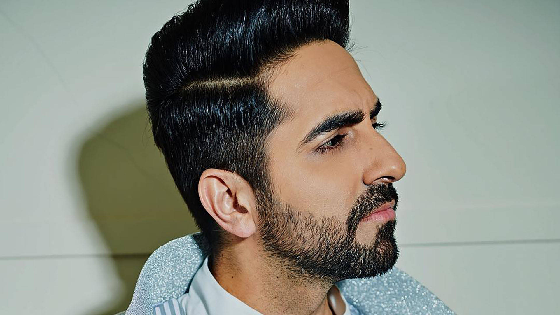 ayushmann khurrana 桌布 壁紙 — 一個擁有時尚蓬巴杜髮型和鬍鬚的男人，靠著素牆若有所思地側視。
