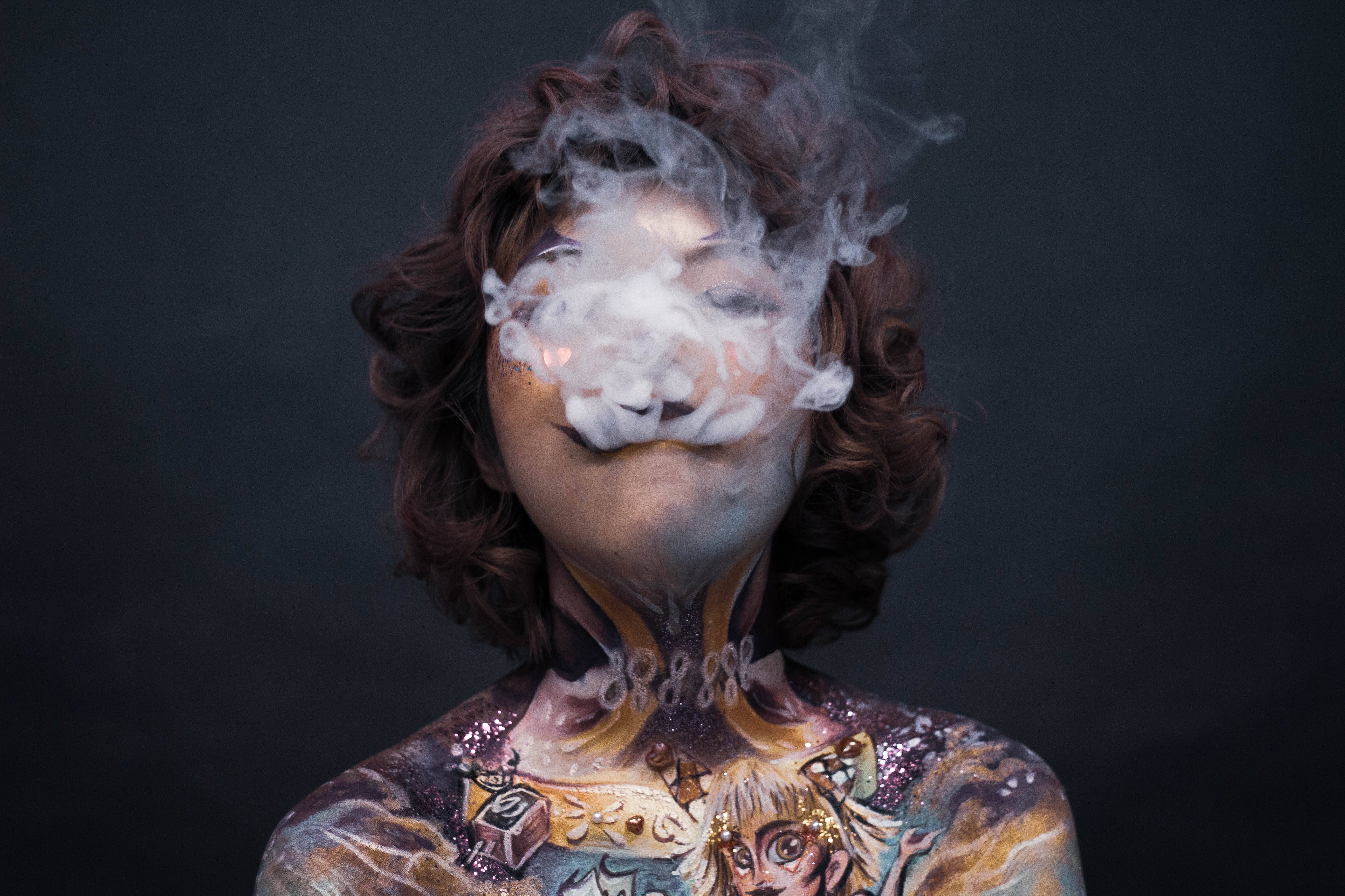 抽菸 桌布 壁紙 — 一個擁有棕色捲髮和複雜身體彩繪的人，在黑暗背景下呼出煙霧，煙霧在他們臉部周圍盤旋。