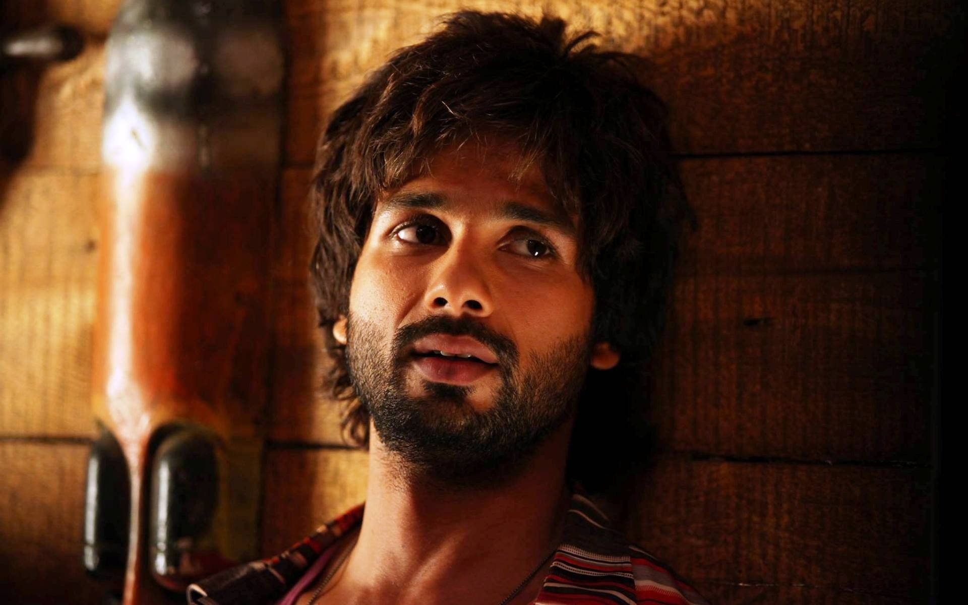 Shahid Kapoor 桌布 壁紙 — 一個擁有深色凌亂頭髮和鬍鬚的男人，在溫暖的木牆上，柔和的金色燈光下向上凝視。