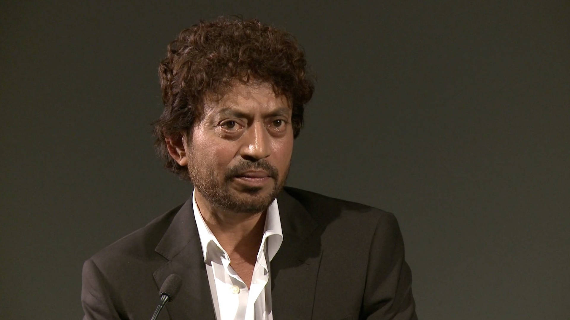 Irrfan Khan 桌布 壁紙 — 一個擁有深色捲髮和鬍鬚的男人，身穿深色西裝內搭白襯衫，對著純灰色背景的麥克風說話。