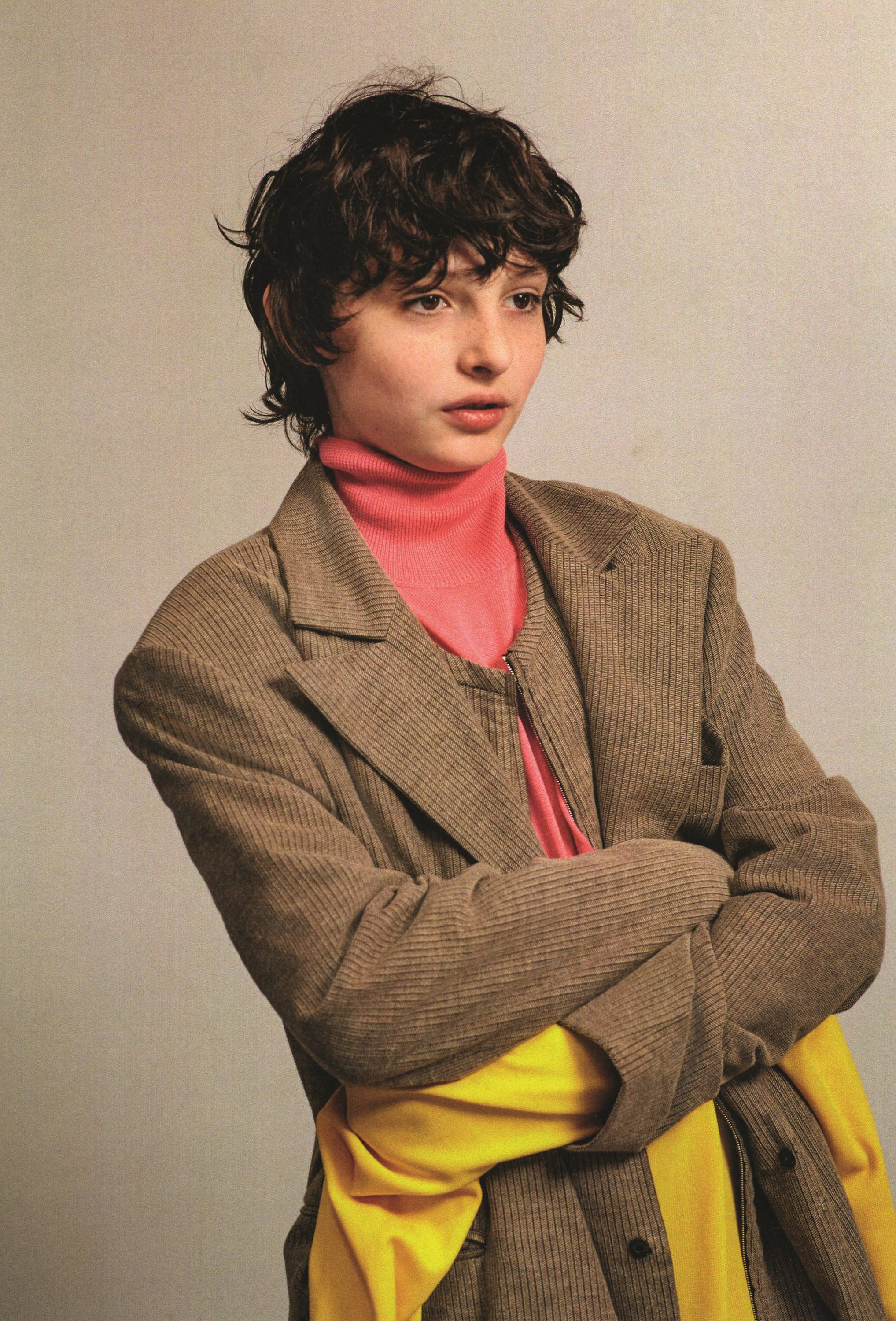 finn wolfhard 桌布 壁紙 — 一個擁有深色捲髮的人穿著棕色燈芯絨西裝外套，內搭粉紅色高領毛衣和黃色褲子，雙手交叉站在純色背景前。