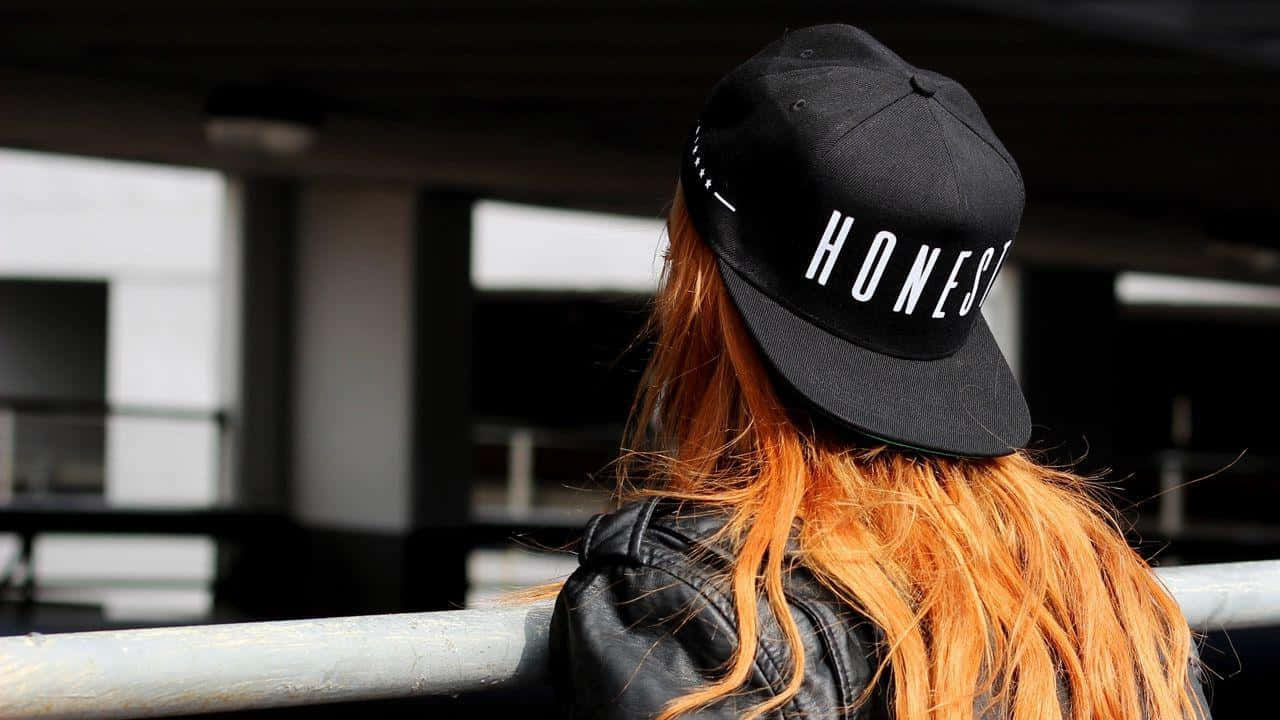 黑色 桌布 壁紙 — 一個擁有火橙色頭髮、戴著黑色「HONEST」帽子的人，在模糊的城市背景下側面站立。