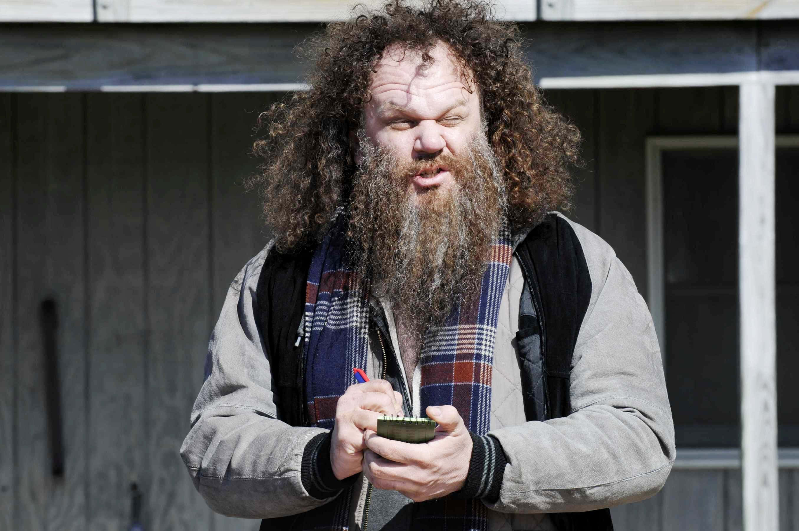 John C. Reilly 桌布 壁紙 — 一個擁有狂野捲髮和長鬍鬚的男人，站在木建築外面，手持一個綠色小碗。