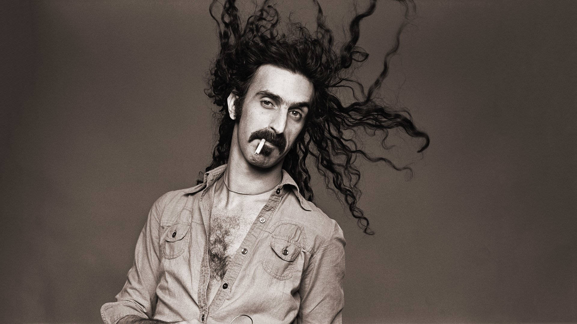 Zappa 桌布 壁紙 — 一個擁有狂野捲髮和鬍子的男人，在純灰色背景前抽煙，呈現戲劇性的黑白肖像。