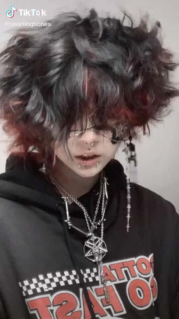 emo 美學 桌布 壁紙 — 一個擁有狂野黑紅髮色及多個穿孔的人，穿著一件帶有粗體紅色文字和層疊銀鏈的黑色連帽衫。