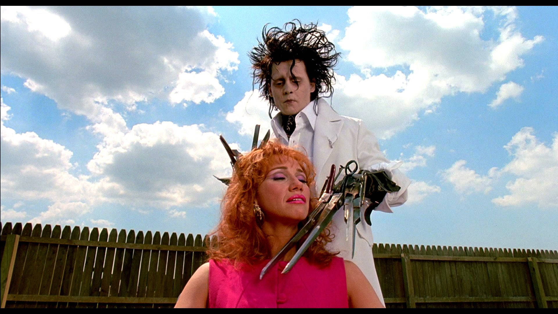 Edward Scissorhands 桌布 壁紙 — 一個擁有狂野黑髮和金屬爪的男人站在一個穿著粉紅色上衣的女人身後，背景是藍天白雲。