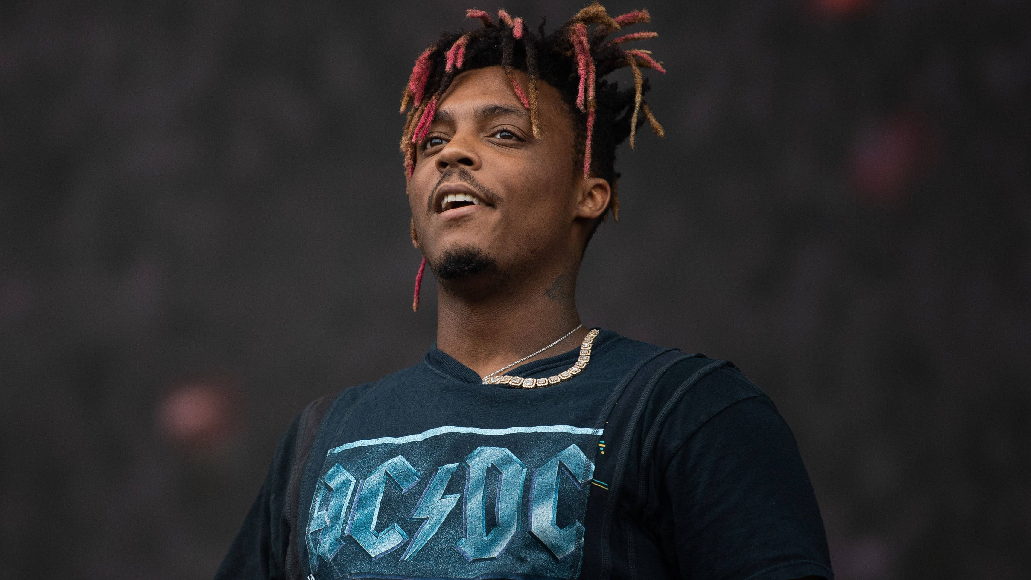 Juice Wrld Desktop 桌布 壁紙 — 一個擁有紅色和棕色辮子的男人，穿著ACDC T恤，在黑暗模糊的背景前向上看，表情豐富。