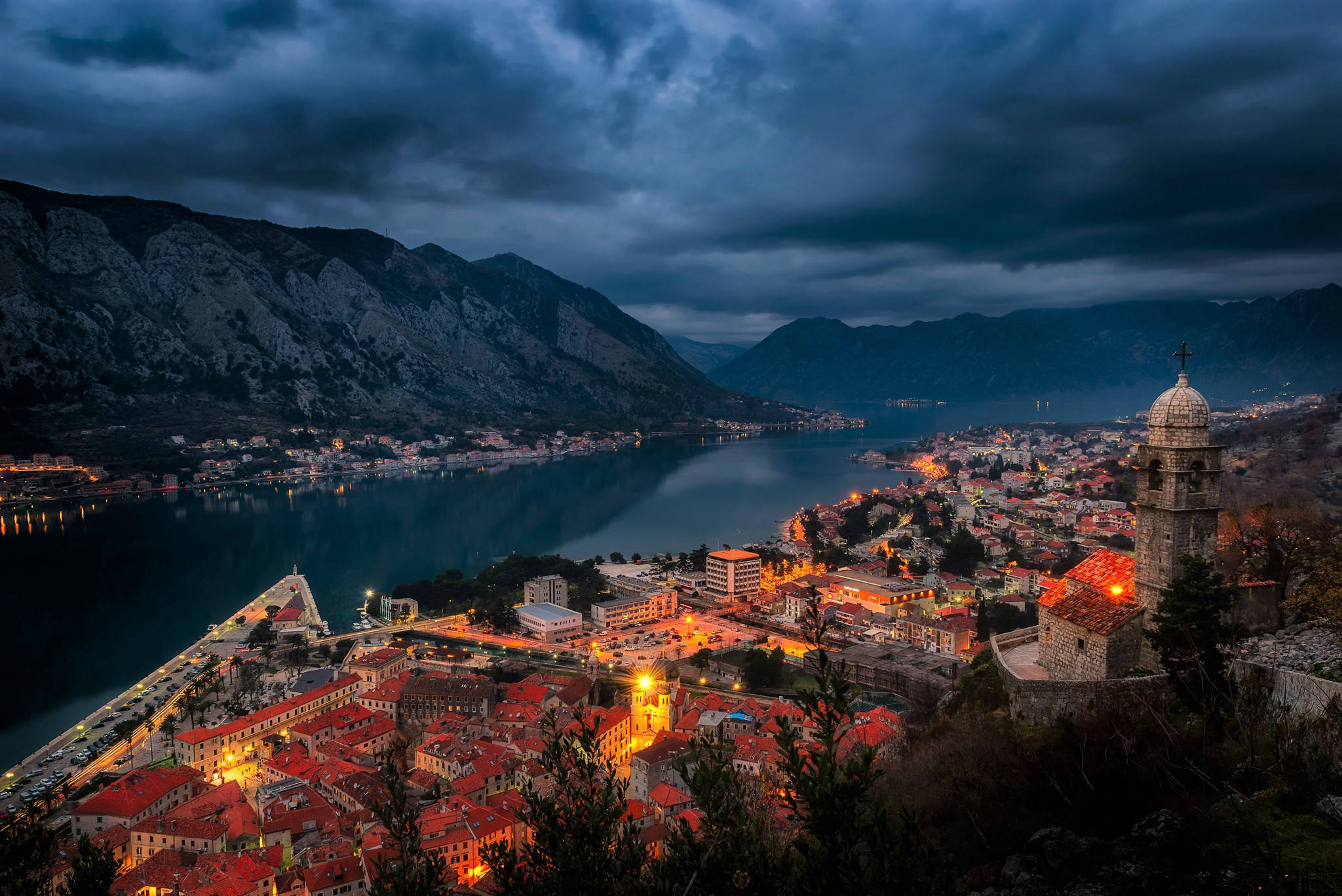 Montenegro 桌布 壁紙 — 一個擁有紅色屋頂的海邊小鎮，在陰暗多雲的天空下，於群山環抱的寧靜海灣旁溫暖地發光。