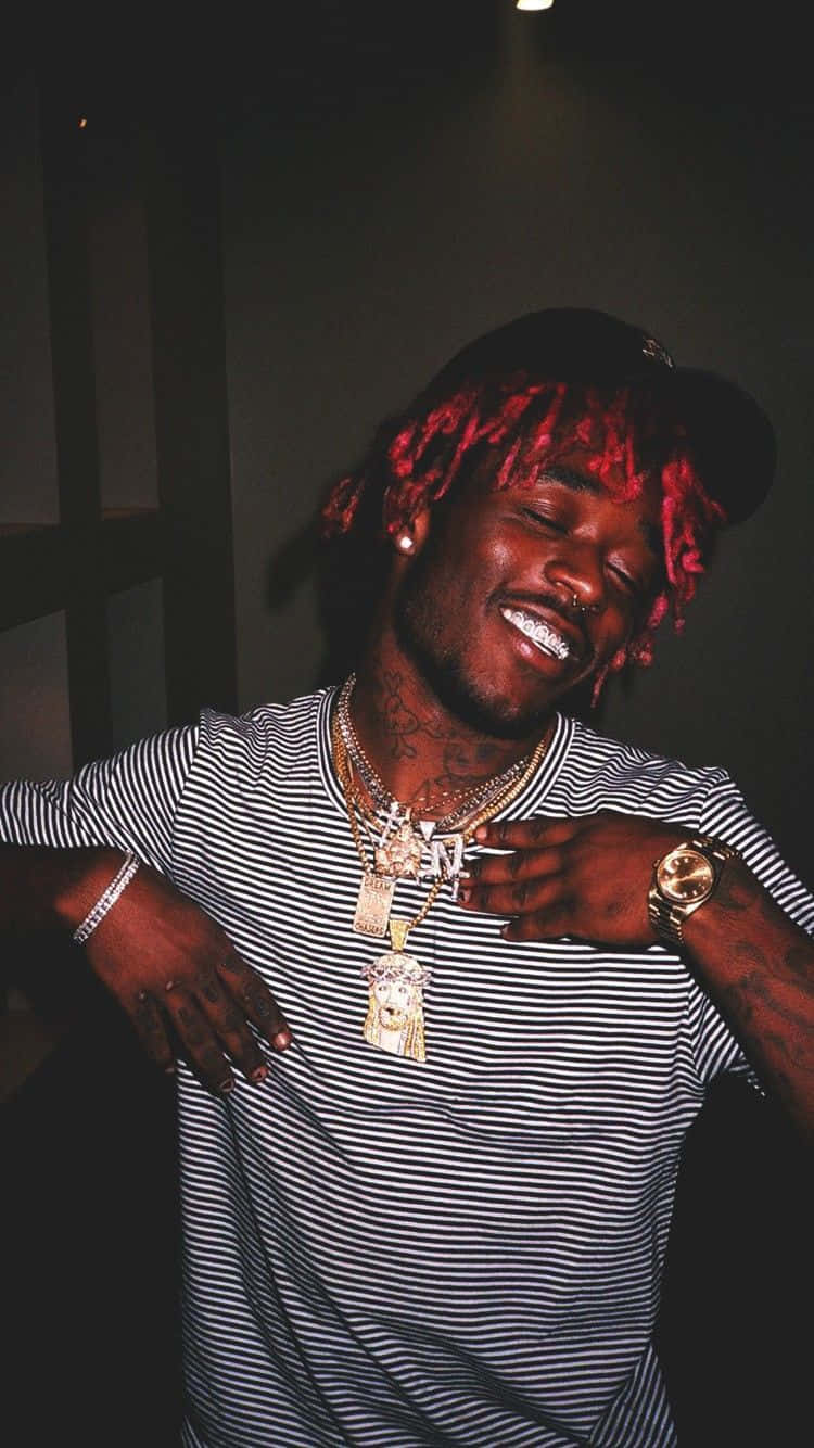 說唱歌手 桌布 壁紙 — 一個擁有紅色辮子頭和金色鏈條的男人，在昏暗的房間裡穿著條紋襯衫自信地微笑。