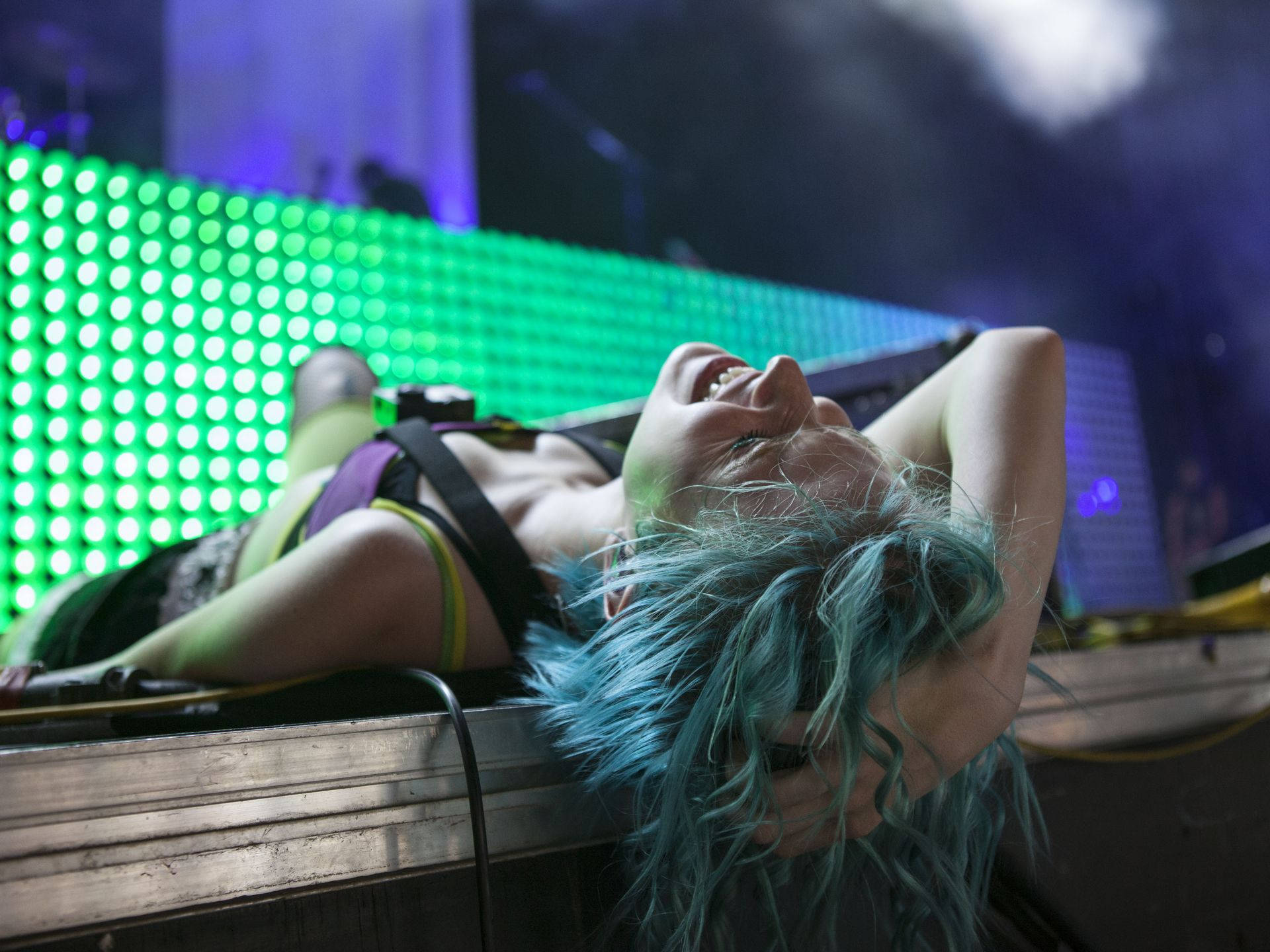 Hayley Williams 桌布 壁紙 — 一個擁有藍綠色頭髮的人躺在舞台上，在充滿活力的綠色和紫色燈光下開懷大笑。