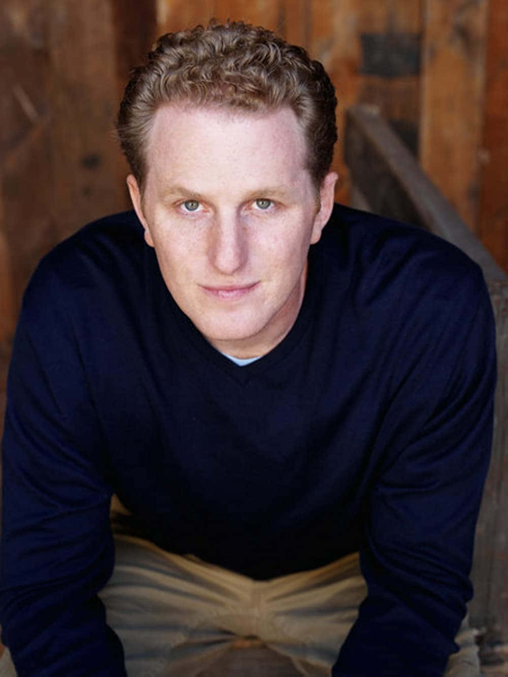 Michael Rapaport 桌布 壁紙 — 一個擁有金色捲髮和藍色眼睛的男人，穿著海軍藍毛衣，在質樸的木質背景前直視鏡頭。