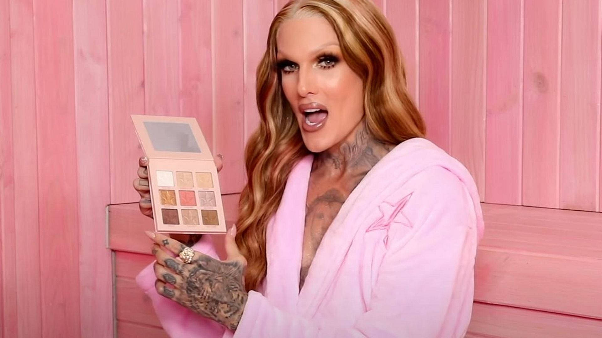Jeffree Star 桌布 壁紙 — 一個擁有金色長髮和刺青的人穿著粉紅色浴袍，手持化妝盤，靠在粉紅色木牆上。