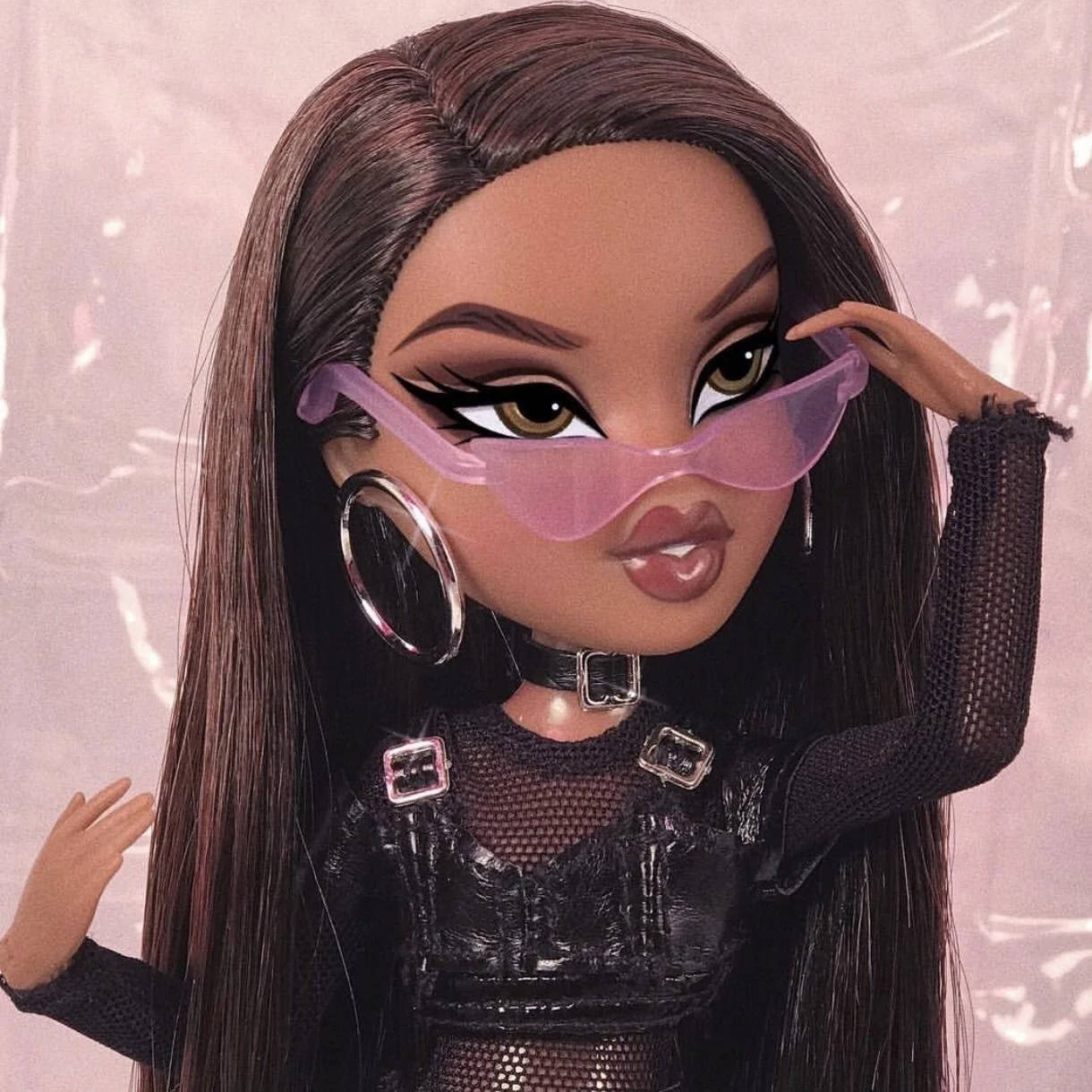 bratz 美學 桌布 壁紙 — 一個擁有長長棕色頭髮和粉紅色太陽眼鏡的 Bratz 公仔，在柔和的粉紅色背景前，自信地穿著黑色網狀服裝擺姿勢。