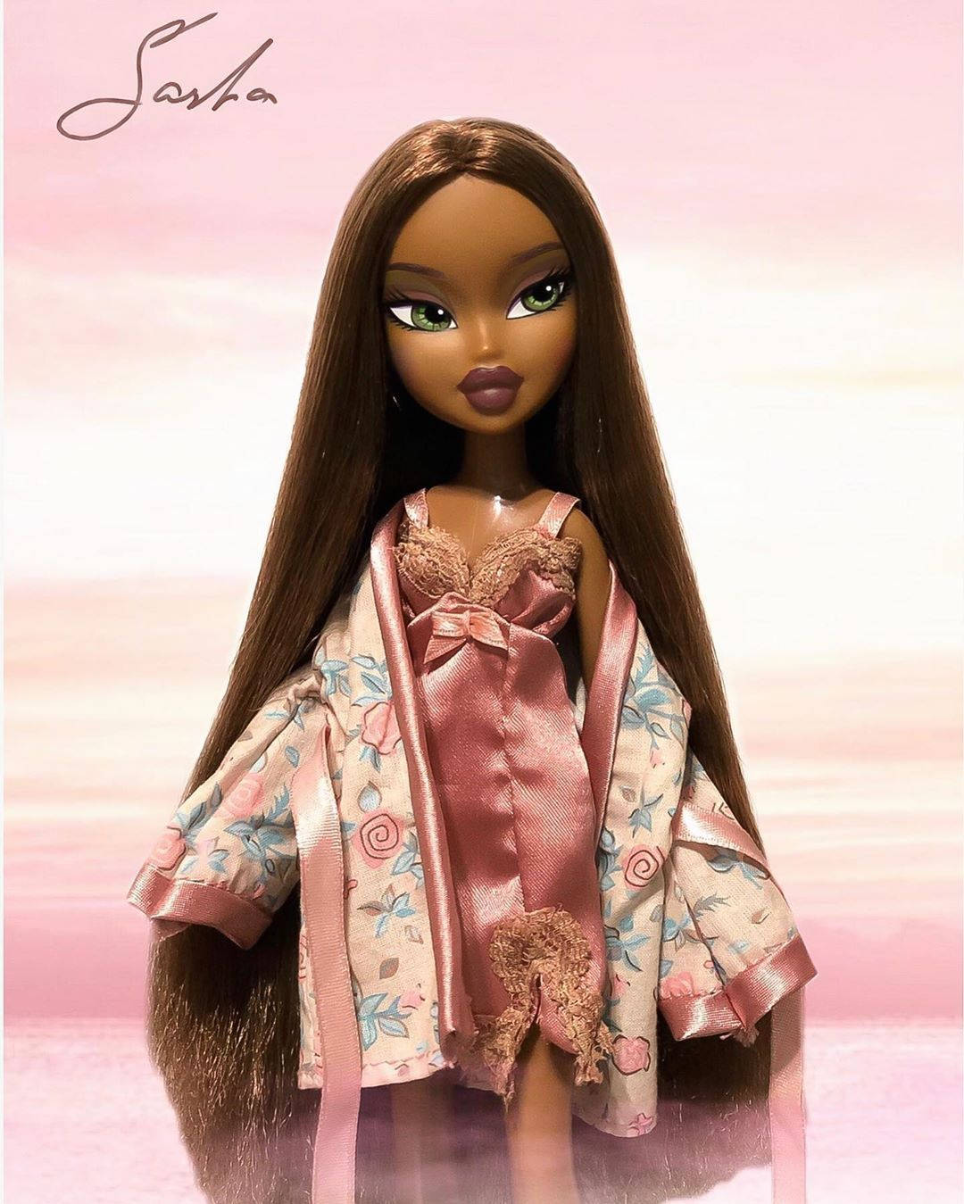 Bratz 桌布 壁紙 — 一個擁有長長棕色頭髮和綠色眼睛的 Bratz 娃娃，身穿粉紅色花卉睡袍，內搭緞面上衣，背景是柔和的粉紅色漸層。