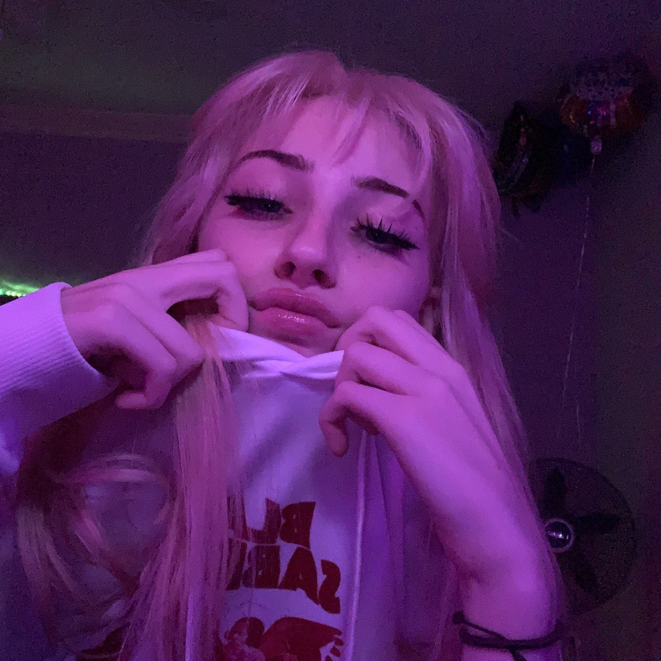 girly pink aesthetic 桌布 壁紙 — 一個擁有長長金髮和戲劇性睫毛的人，在紫色燈光下拉扯白色連帽衫的領口。