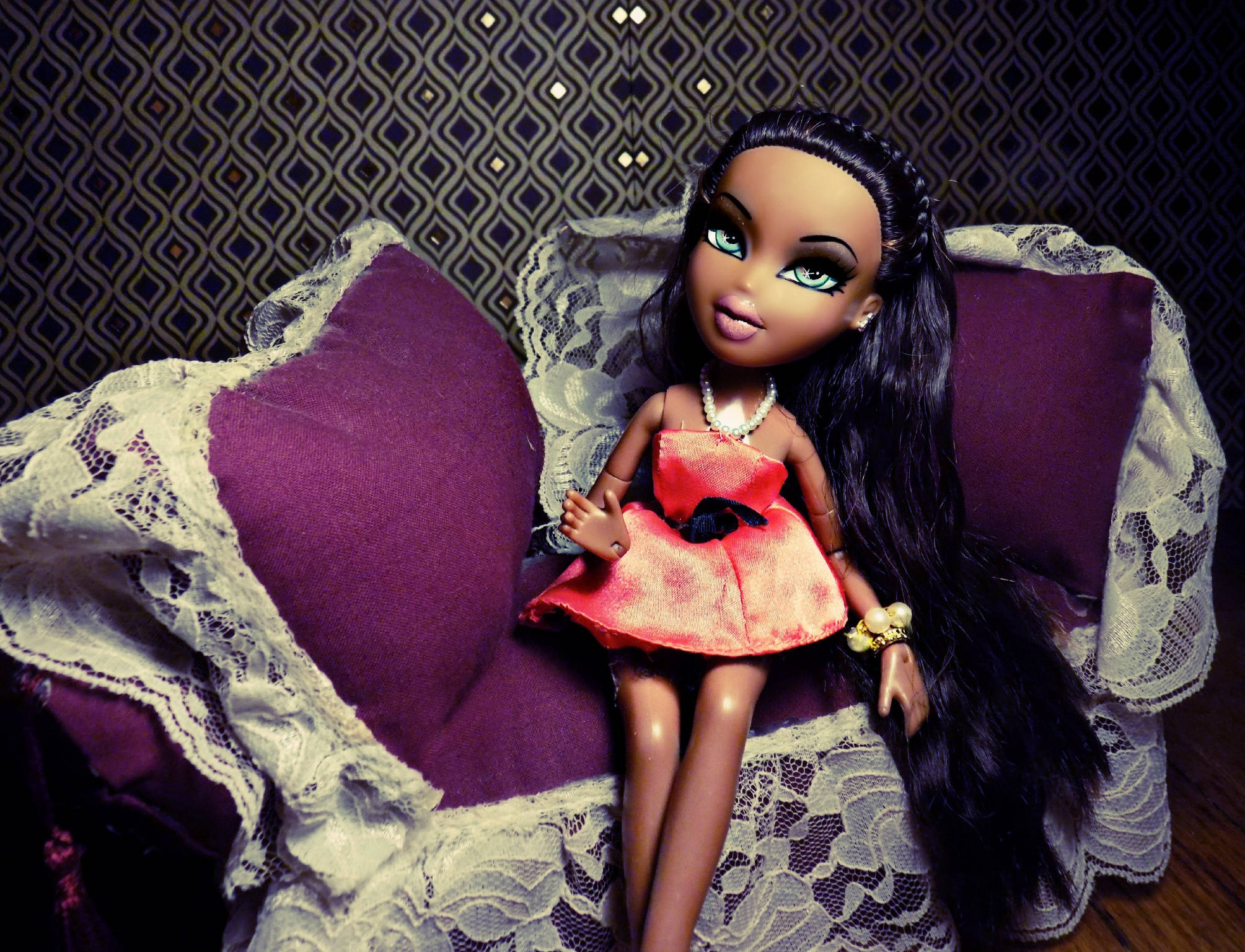 bratz 美學 桌布 壁紙 — 一個擁有長長黑髮和發光綠色眼睛的洋娃娃優雅地坐在一張飾有白色蕾絲的紫色軟墊椅子上，背景是圖案壁紙。