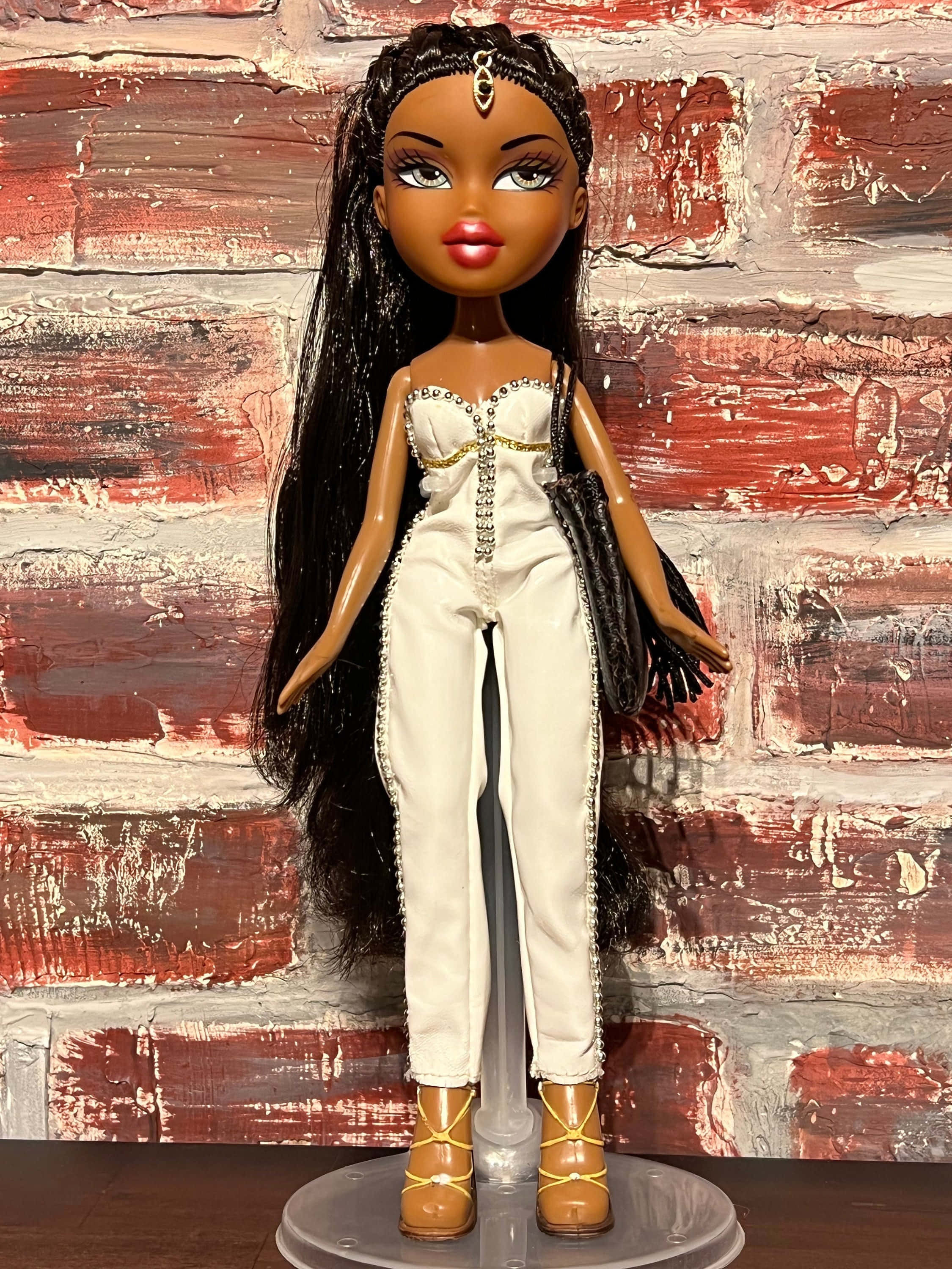 bratz 美學 桌布 壁紙 — 一個擁有長長黑髮和金色配飾的 Bratz 公仔，站在風化的紅磚牆前，穿著白色連身褲和棕色靴子。