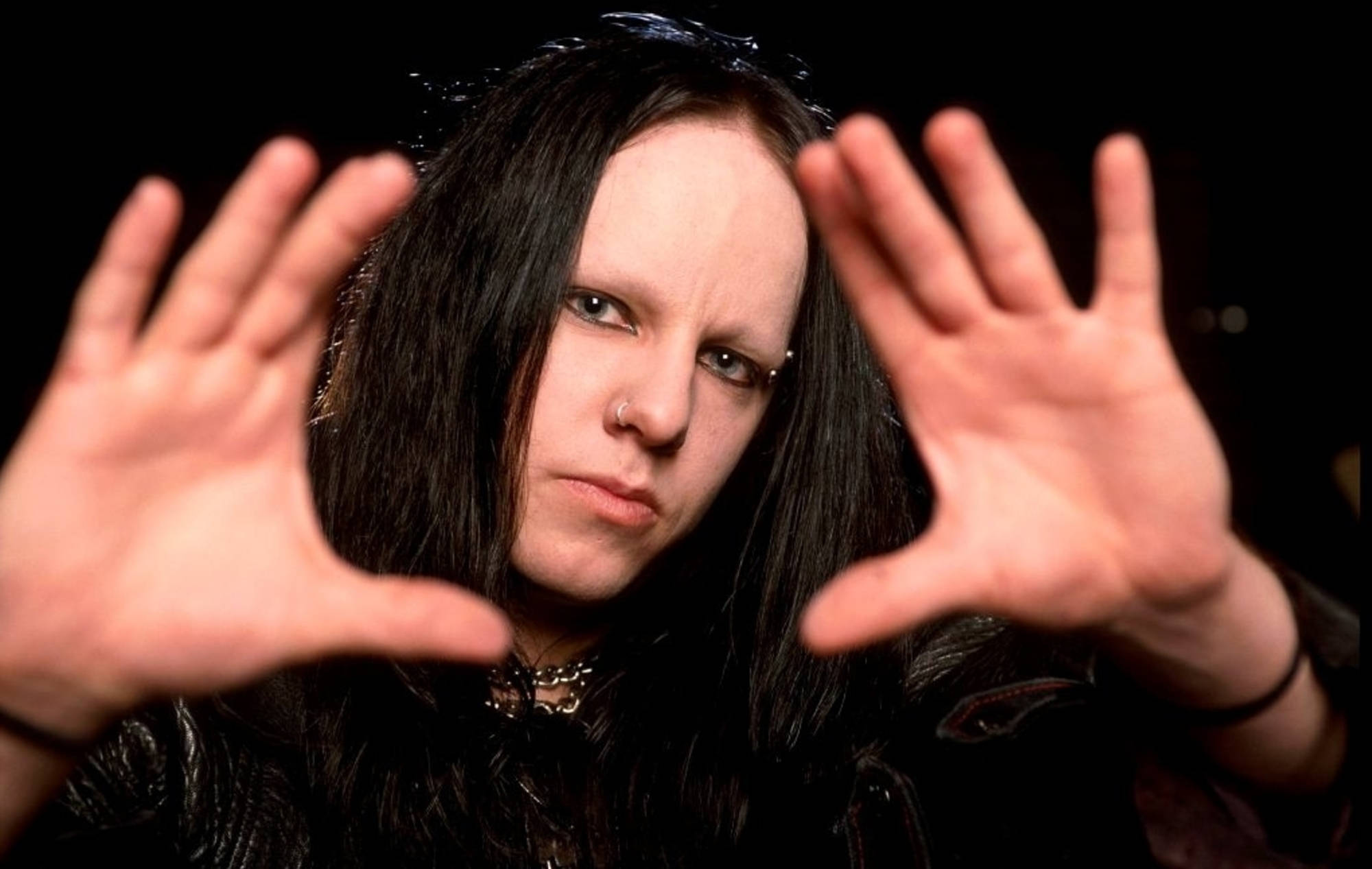 Joey Jordison 桌布 壁紙 — 一個擁有長黑髮和鼻環的人，雙手向前伸出，在黑色背景前表情嚴肅。