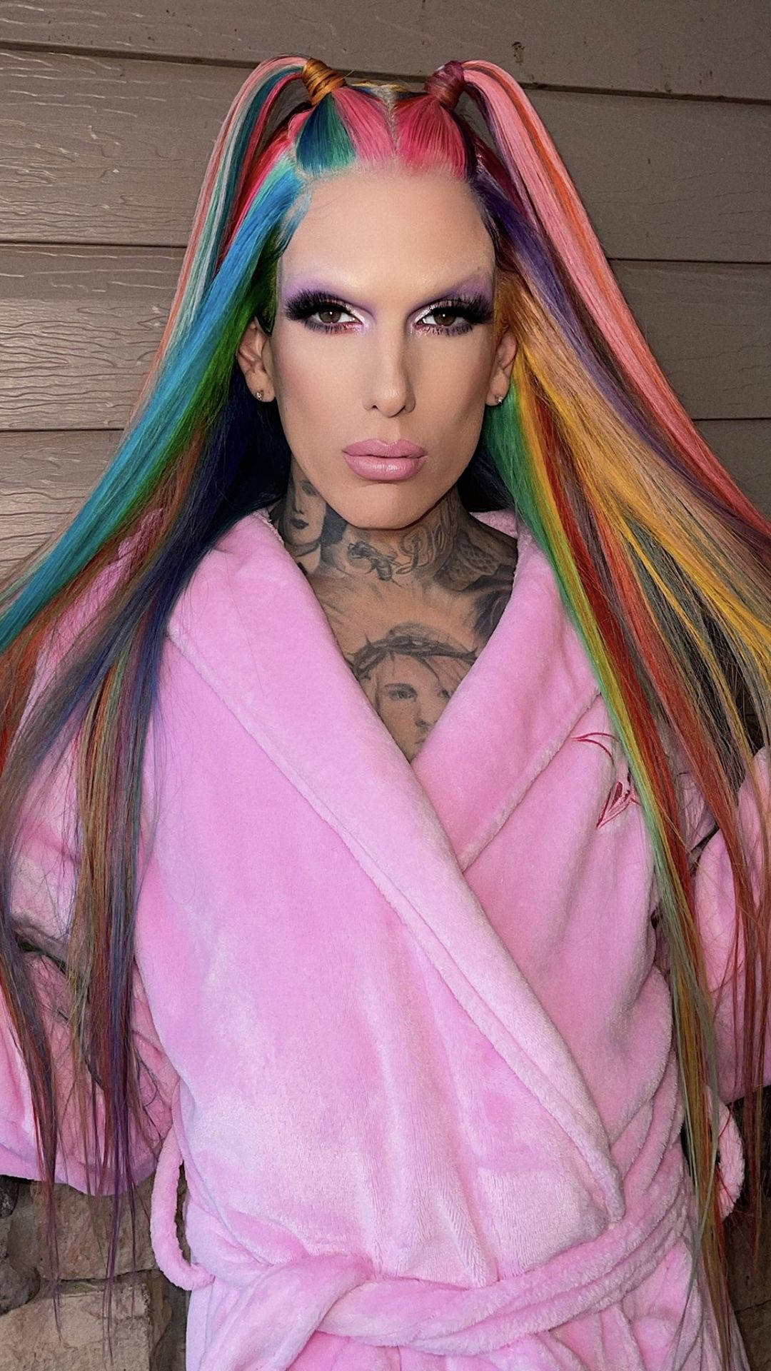 Jeffree Star 桌布 壁紙 — 一個擁有鮮豔彩虹色辮子頭髮和濃妝的人，穿著毛茸茸的粉紅色浴袍，靠著木牆。