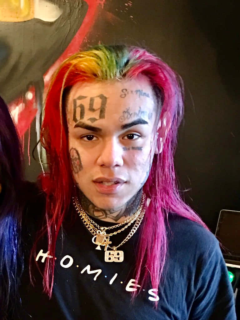 69 Rapper 桌布 壁紙 — 一個擁有鮮豔彩虹髮色和臉部刺青的人，穿著黑色 'HOMIES' 襯衫直視鏡頭。