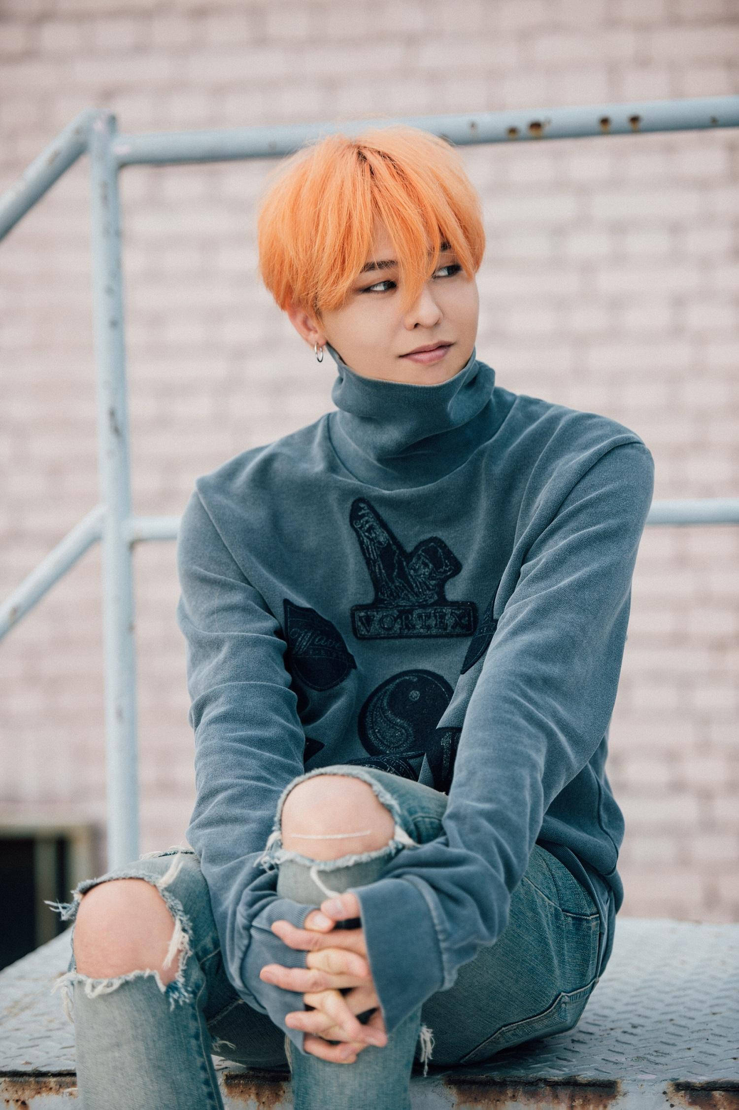 G-dragon 桌布 壁紙 — 一個擁有鮮豔橙色頭髮的人，穿著灰色高領毛衣和破洞牛仔褲，坐在金屬屋頂上，背景是磚牆，若有所思。