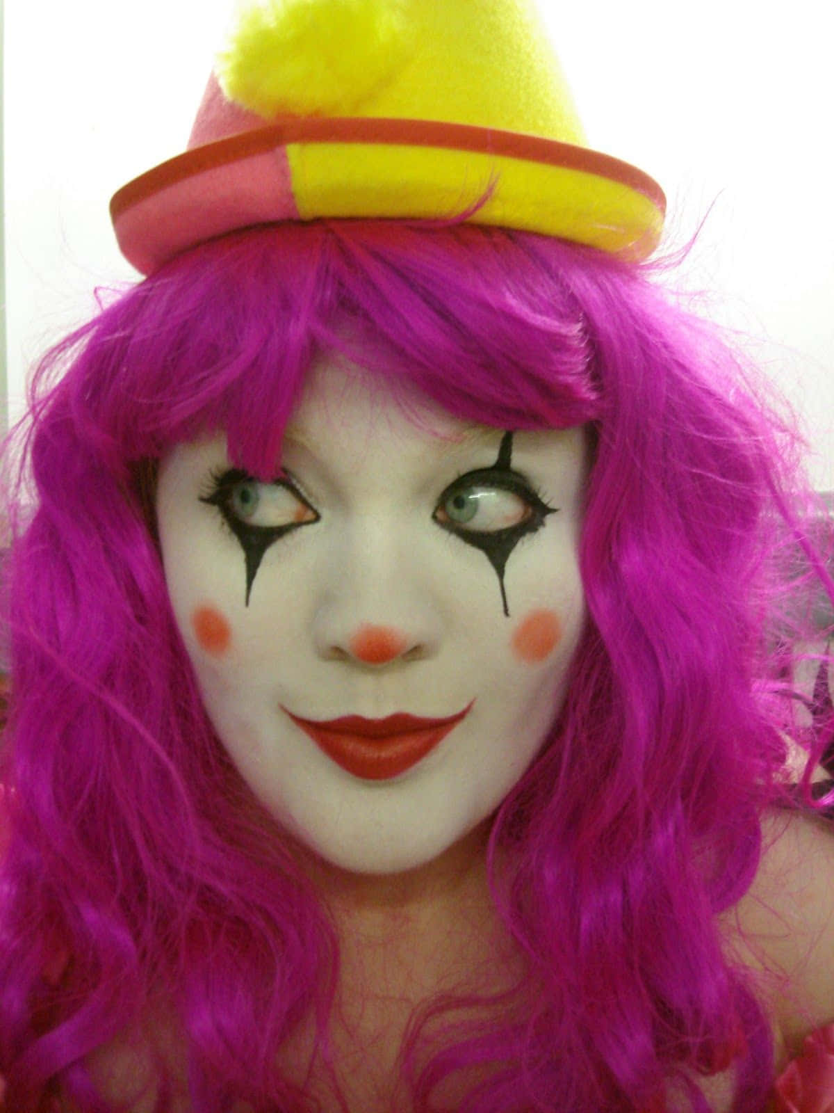 搞笑 桌布 壁紙 — 一個擁有鮮豔粉紅色頭髮和小丑妝容的人，在純色背景前戴著一頂色彩繽紛的帽子。