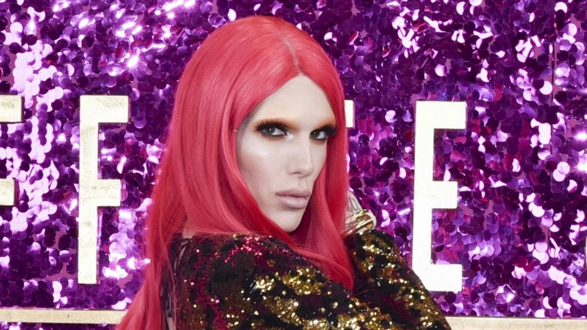 Jeffree Star 桌布 壁紙 — 一個擁有鮮豔紅髮和大膽妝容的人，在帶有白色字母的閃亮紫色亮片背景前擺姿勢。