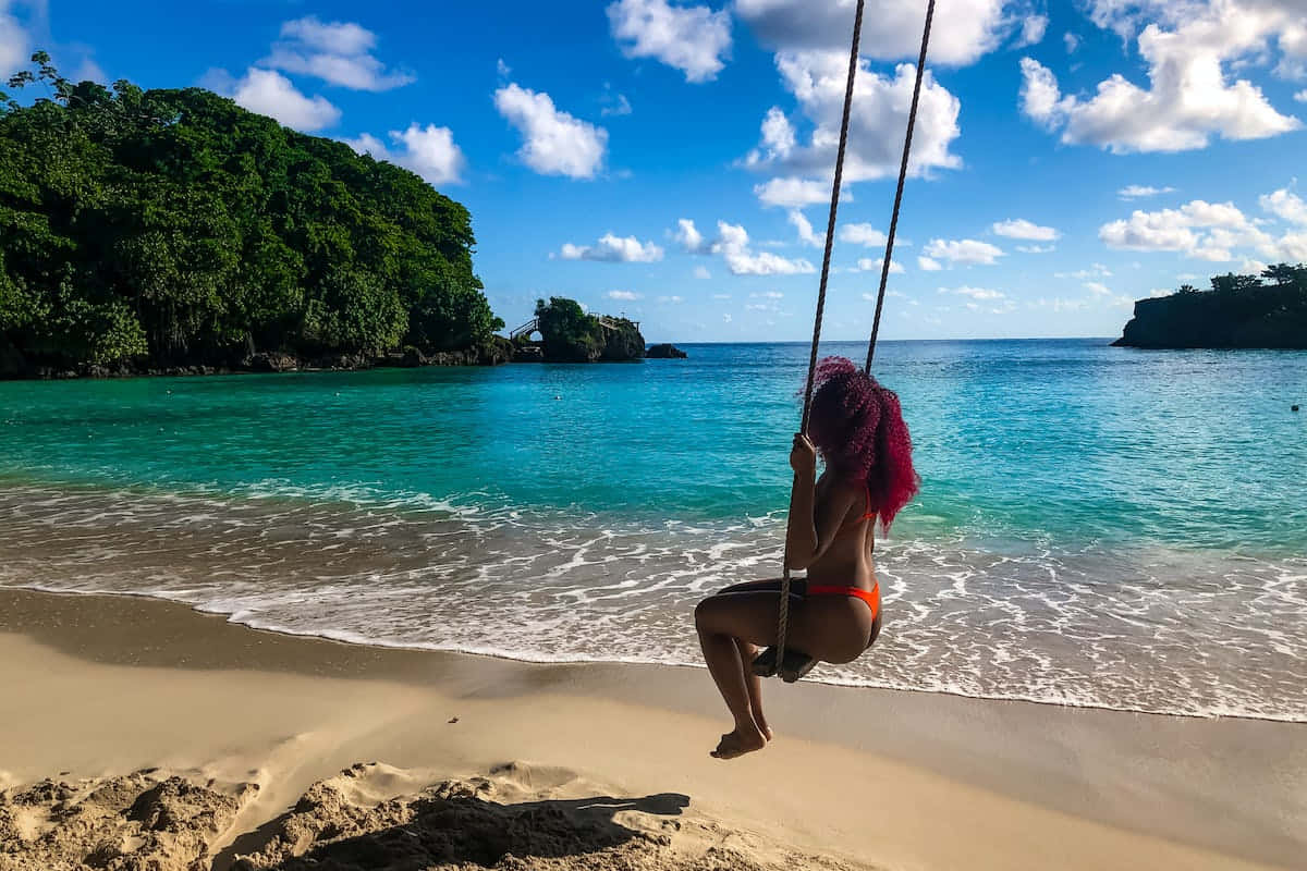 Jamaican Island 桌布 壁紙 — 一個擁有鮮豔紅髮的人，在陽光明媚的沙灘上，用繩索鞦韆在碧綠的海水和茂密的綠色懸崖下，在明亮的藍天下搖擺。