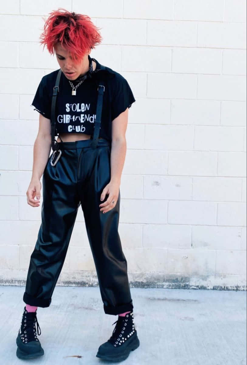 emo 美學 桌布 壁紙 — 一個擁有鮮豔紅髮的人，穿著黑色吊帶褲和閃亮褲子，背景是白色磚牆。