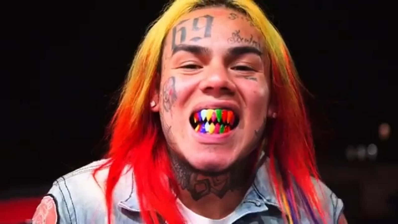 69 Rapper 桌布 壁紙 — 一個擁有鮮豔紅黃色頭髮和彩色牙齒的人，在深色背景下咧嘴大笑。