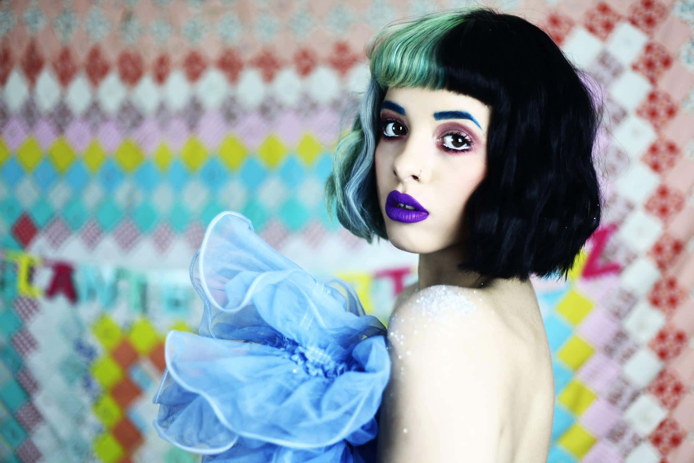 Melanie Martinez Aesthetic 桌布 壁紙 — 一個擁有鮮豔紫色嘴唇和綠色髮尾的黑色鮑伯頭人物，在色彩繽紛的拼布被子背景前擺姿勢。