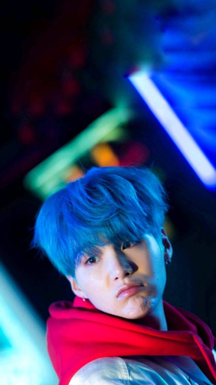 BTS 鎖定螢幕 桌布 壁紙 — 一個擁有鮮豔藍色頭髮和紅色連帽衫的年輕男子，在霓虹燈照亮的黑暗背景下凝視著。