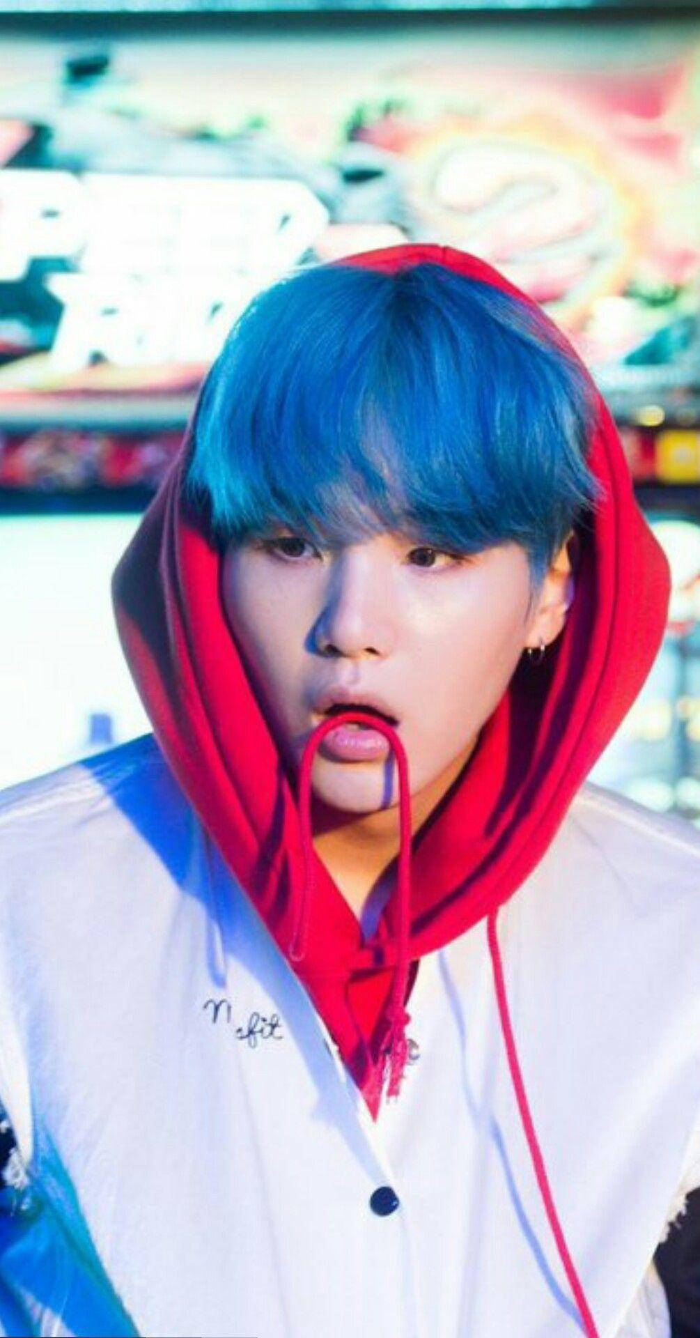 Suga Bts 桌布 壁紙 — 一個擁有鮮豔藍色頭髮的人，穿著紅色連帽衫和白色外套，背景是色彩繽紛且模糊的。