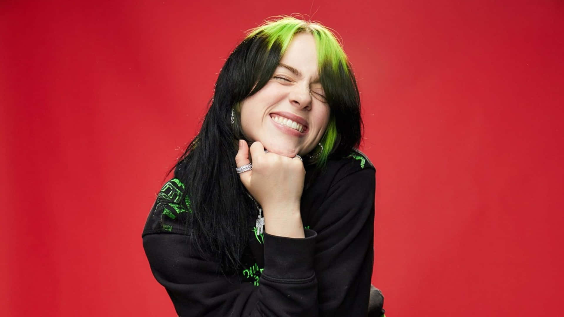 Billie Eilish Laptop 桌布 壁紙 — 一個擁有黑色和螢光綠色頭髮的人，在鮮紅色的背景前，穿著帶有綠色刺繡的黑色連帽衫，開懷大笑。