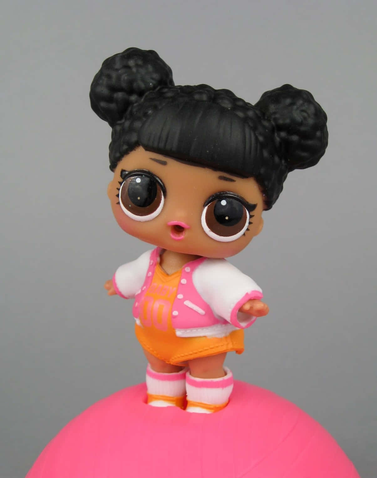 Lol Doll 桌布 壁紙 — 一個擁有黑色捲髮雙馬尾和大眼睛的LOL Surprise娃娃，站在灰色背景前的亮粉紅色球上。