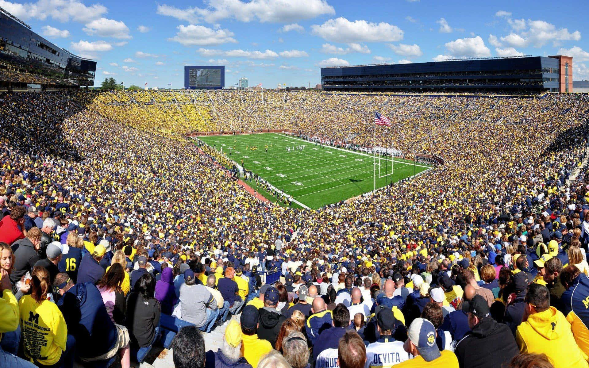 Michigan Football 桌布 壁紙 — 一個擠滿人的足球場，在明亮的藍天下，身穿黃色和藍色的球迷正在為比賽歡呼。