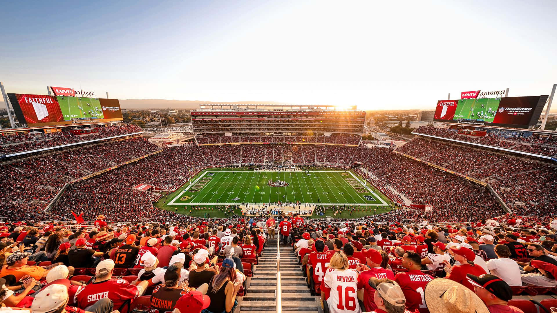 49ers 桌布 壁紙 — 一個擠滿人的足球場沐浴在金色的夕陽下，身穿紅色球衣的球迷在看台上歡呼。