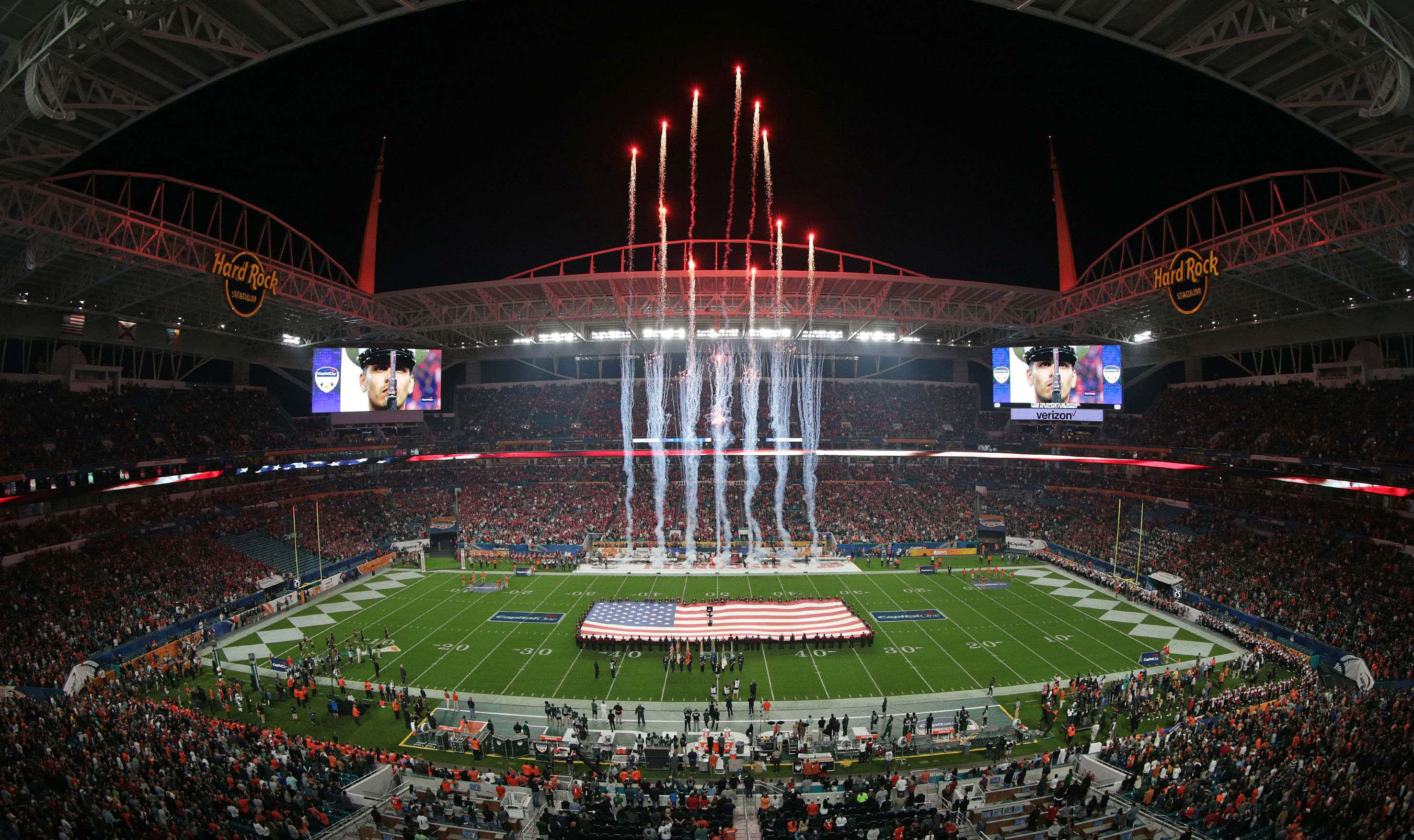 Super Bowl 桌布 壁紙 — 一個擠滿人的體育場在夜晚爆發紅色煙火，上方是一個展示巨型美國國旗的足球場。